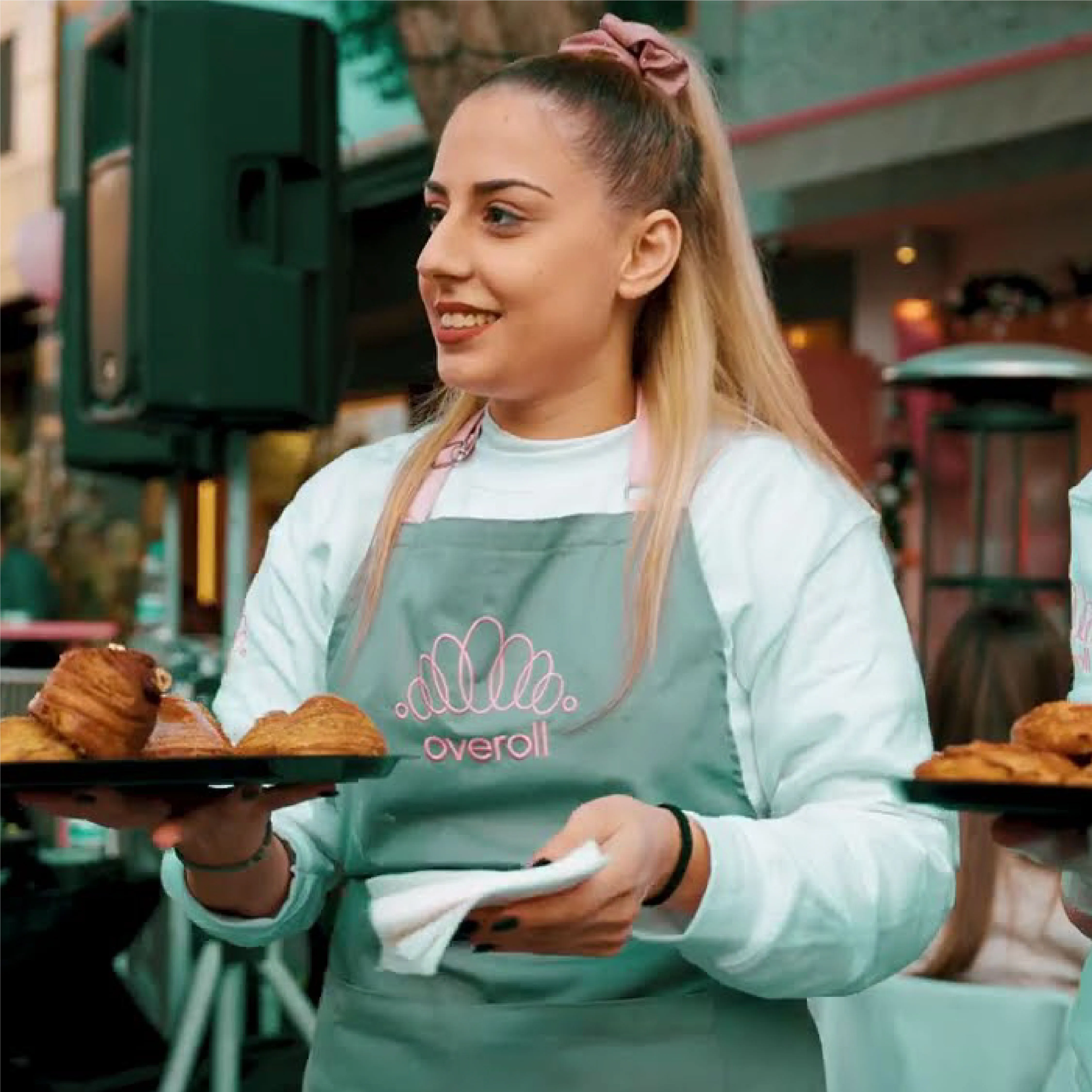 Smiling woman with long blonde hair tied with a scrunchie serving pastries on trays while wearing a green apron with a pink logo.