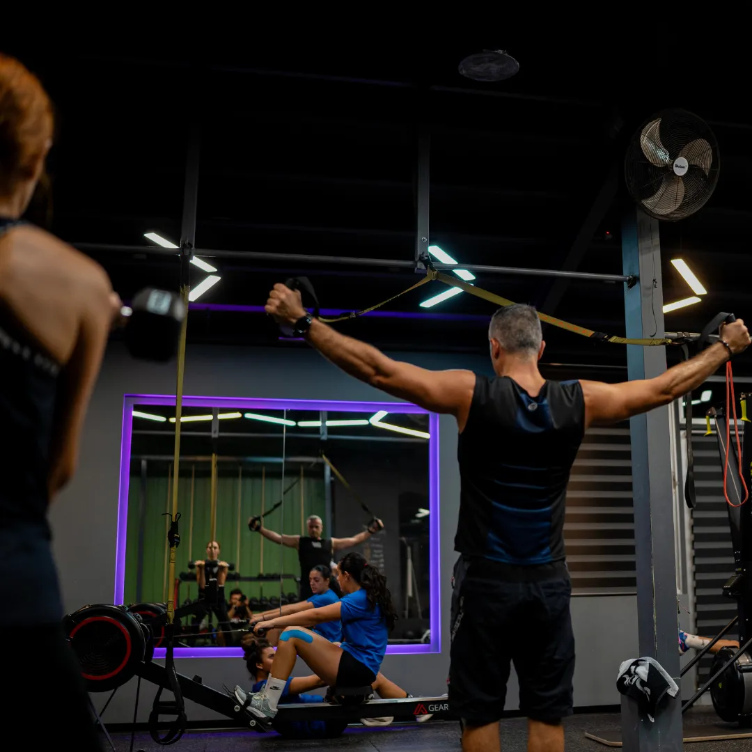 People exercising in a gym with resistance bands and rowing machines, seen in front of a large mirror with purple LED lighting.