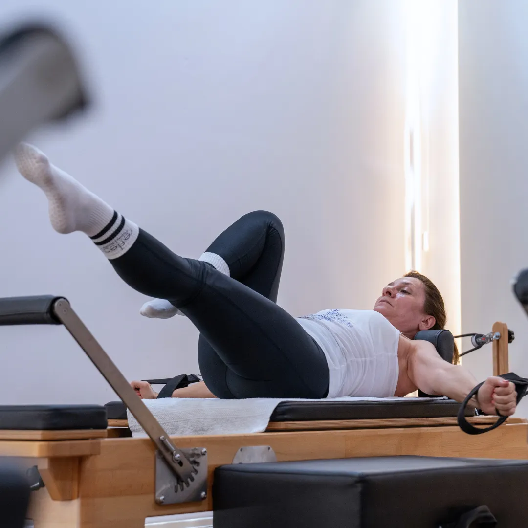 Woman exercising on a Pilates reformer machine, lying on her back with arms extended holding resistance straps and one leg raised.
