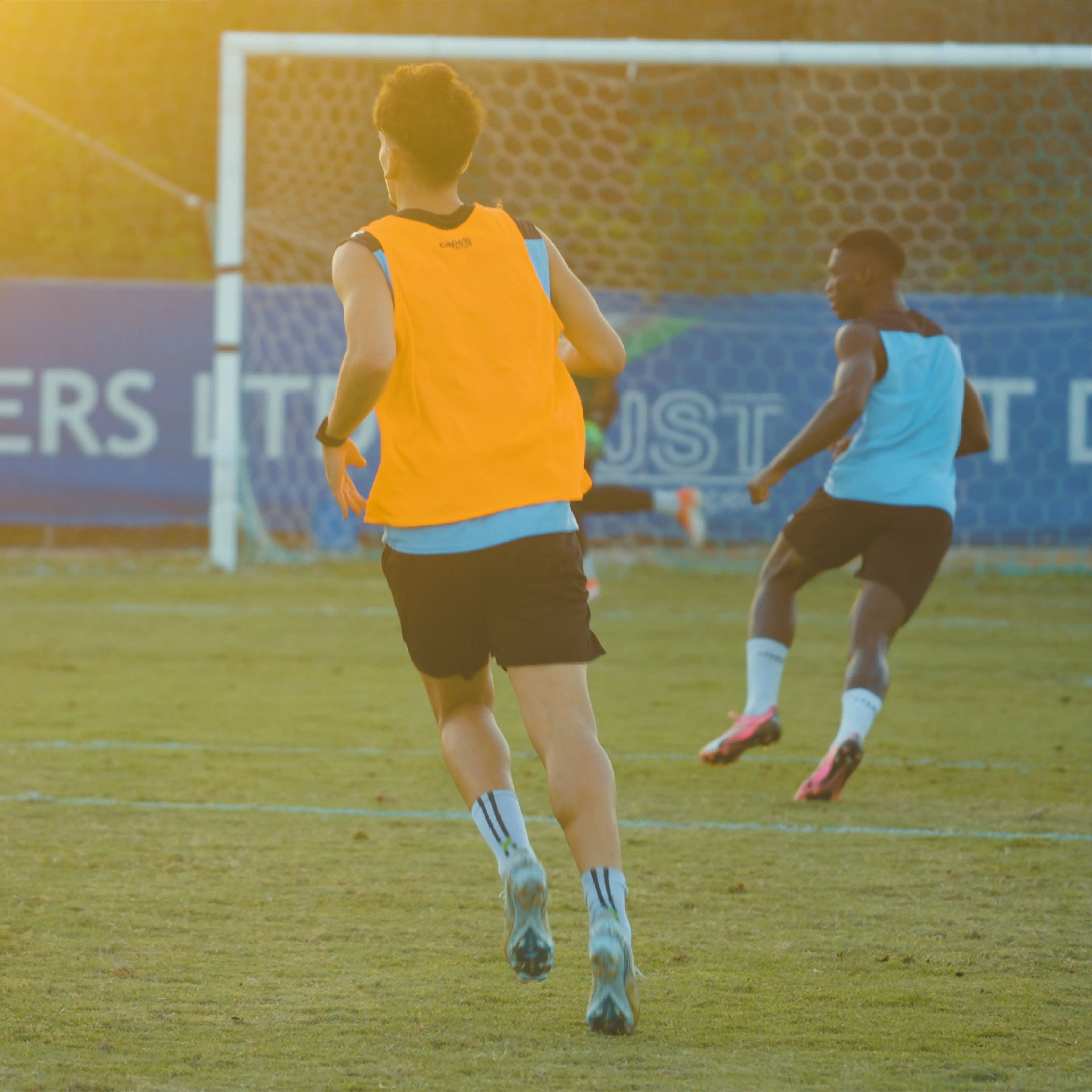 Two soccer players in training gear running on a grassy field toward a goal during sunset.
