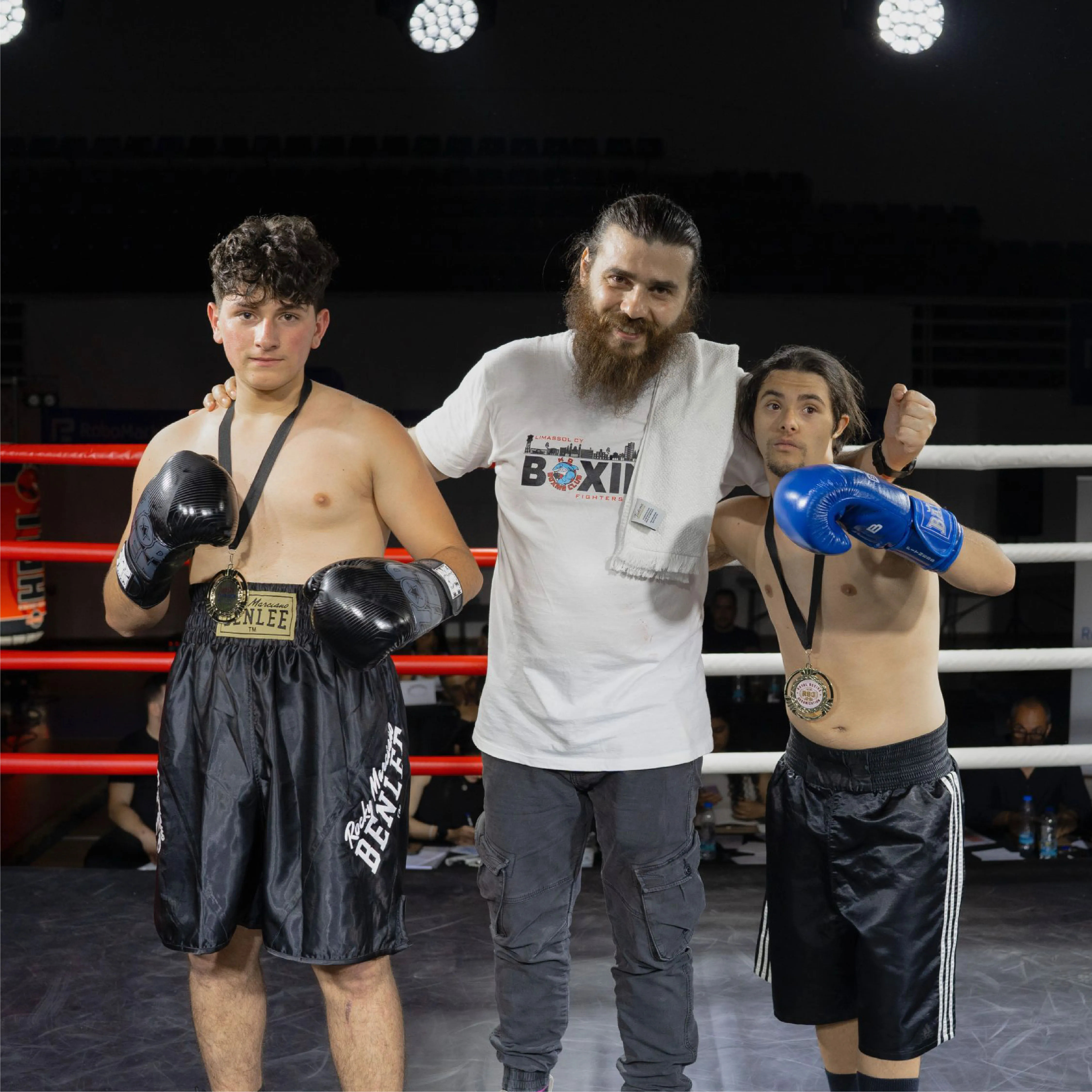 Two young boxers wearing medals and gloves standing beside a man with a beard and towel in a boxing ring.