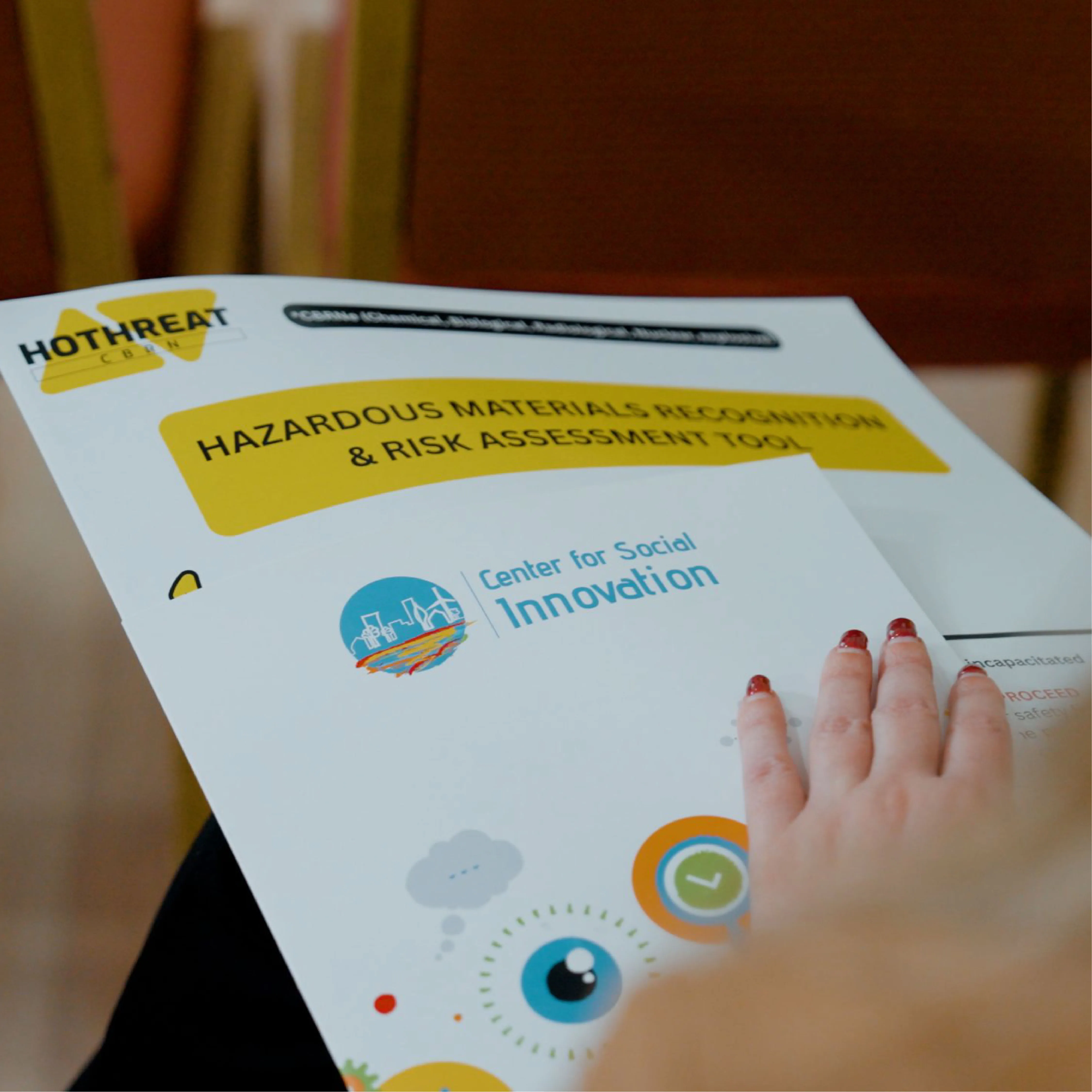 A hand with red-painted nails resting on papers titled Hazardous Materials Recognition & Risk Assessment Tool and Center for Social Innovation.
