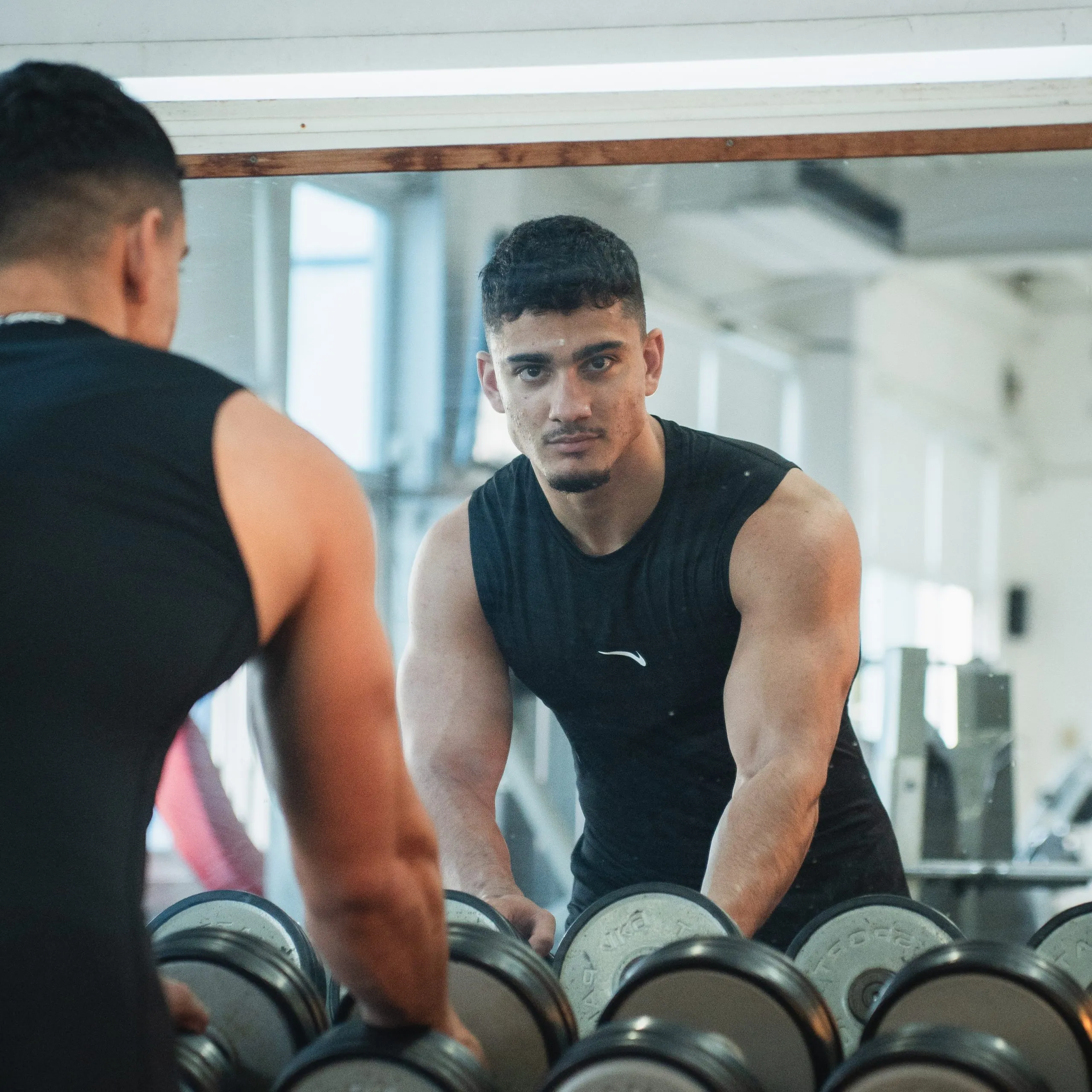 Muscular man in a black sleeveless shirt looking at himself in a gym mirror while holding dumbbells.
