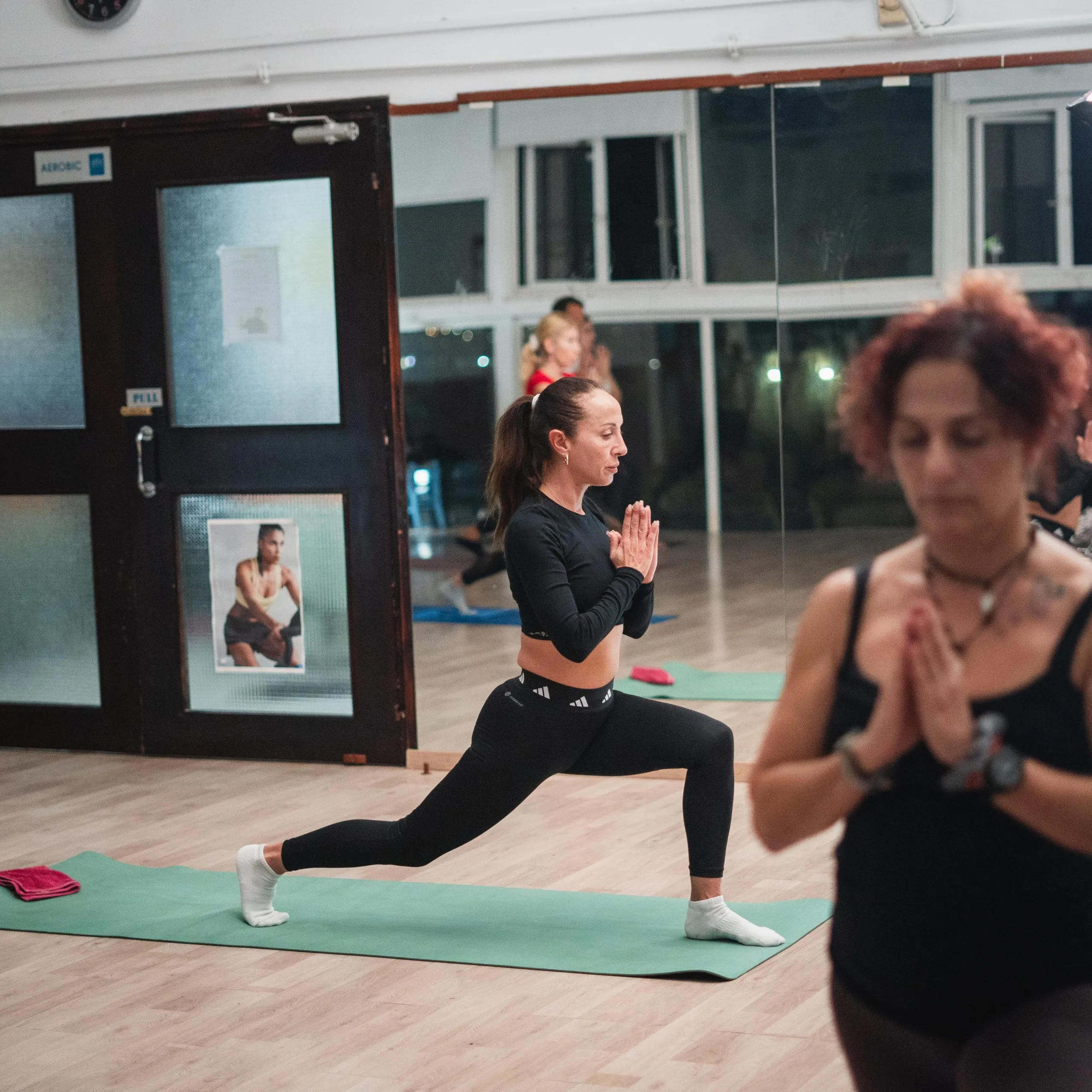 Women practicing yoga indoors on mats, one in a lunge pose with hands in prayer position.