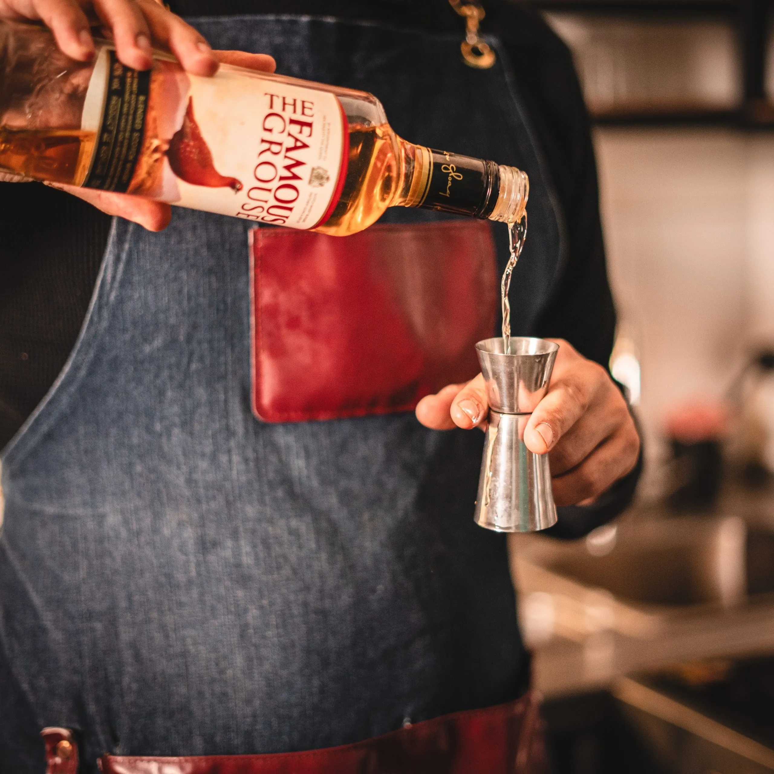 Person wearing a denim apron pouring The Famous Grouse whiskey into a metal jigger.