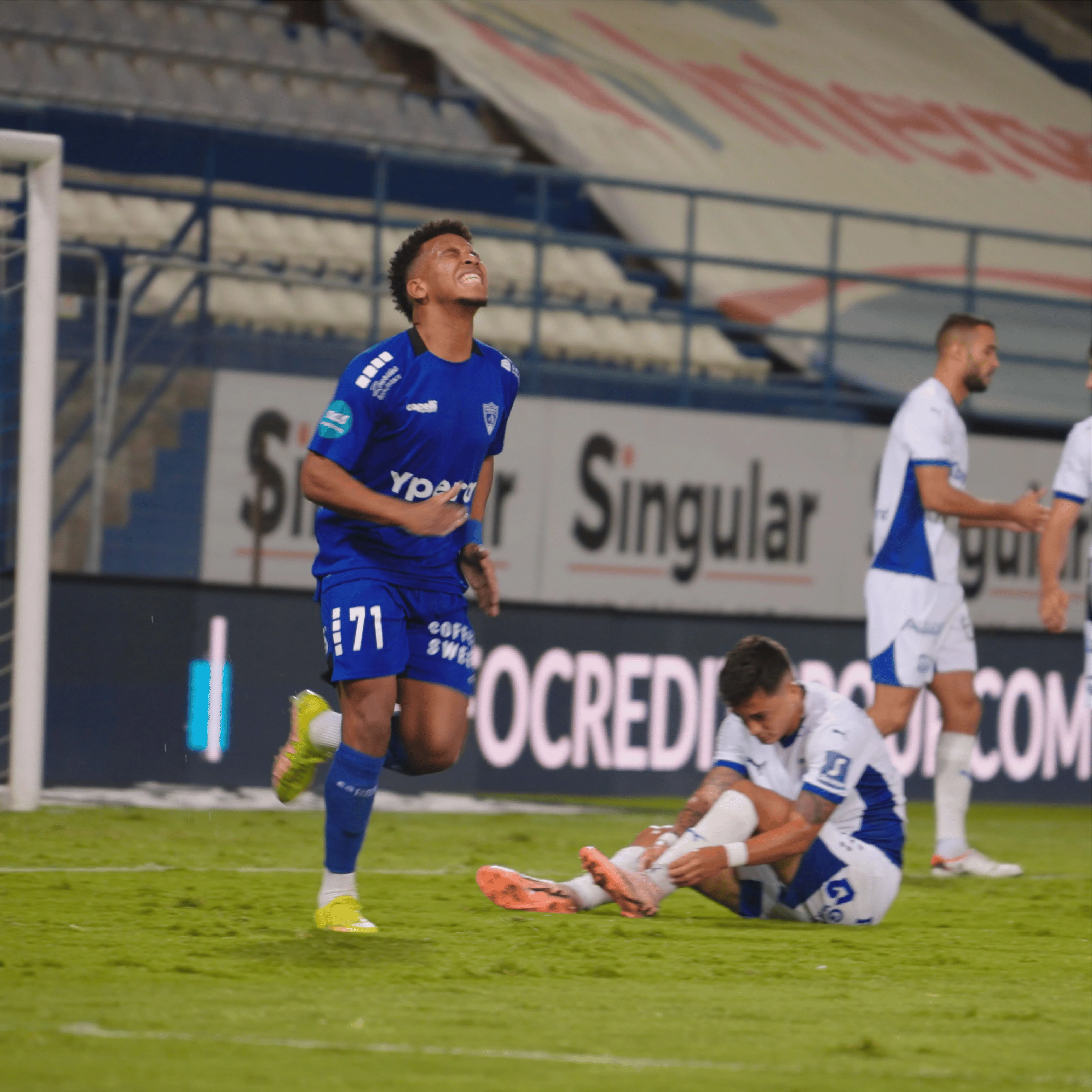 Soccer player in blue jersey showing frustration with clenched fists while another player in white sits on the grass holding his ankle.