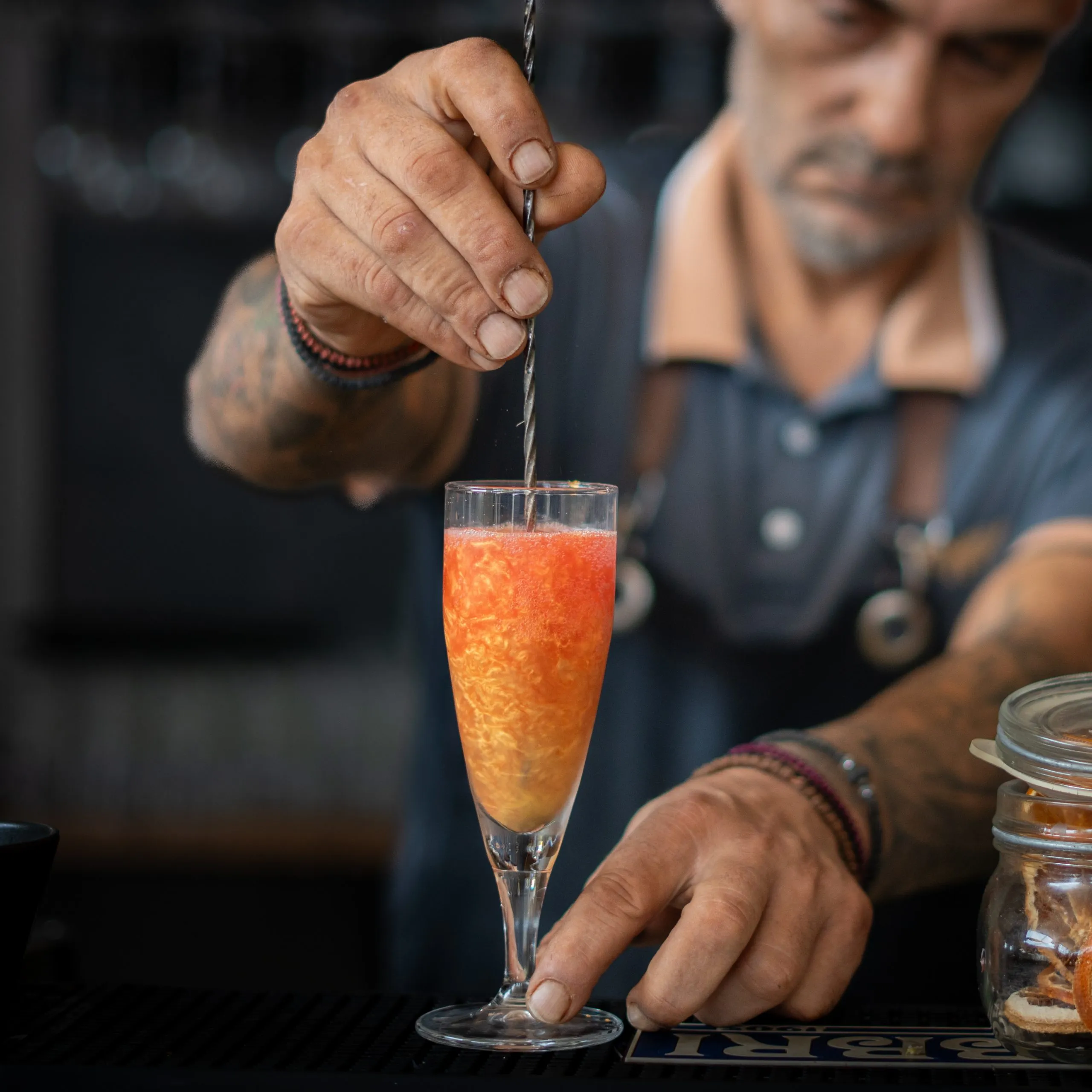 Bartender stirring a layered red and orange cocktail in a champagne flute using a twisted bar spoon.