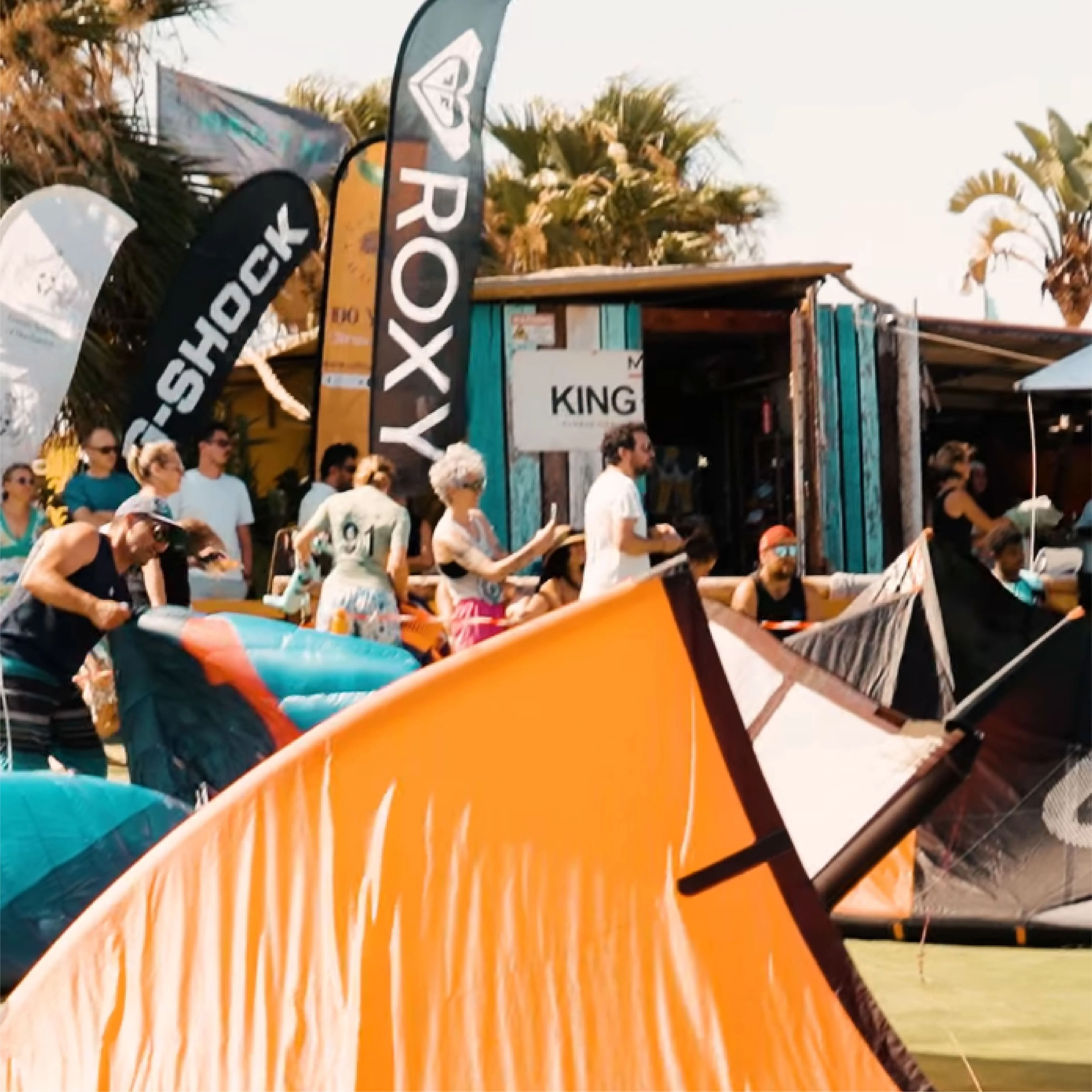 People gathered outdoors around colorful kite surfing equipment under palm trees and branded flags.