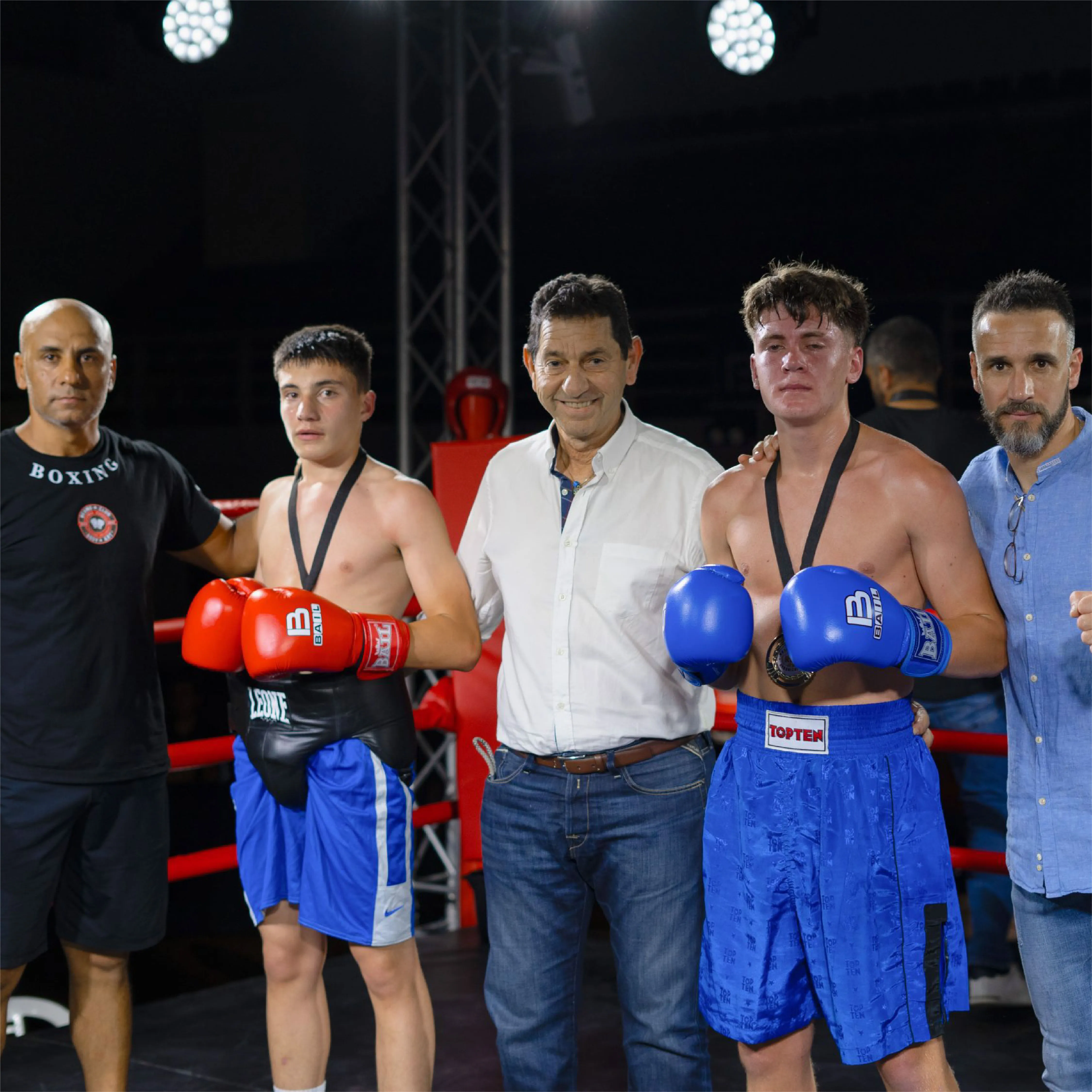 Two young male boxers wearing gloves and medals stand in a boxing ring with two men on each side, one wearing a 'BOXING' t-shirt, and the others in casual shirts.
