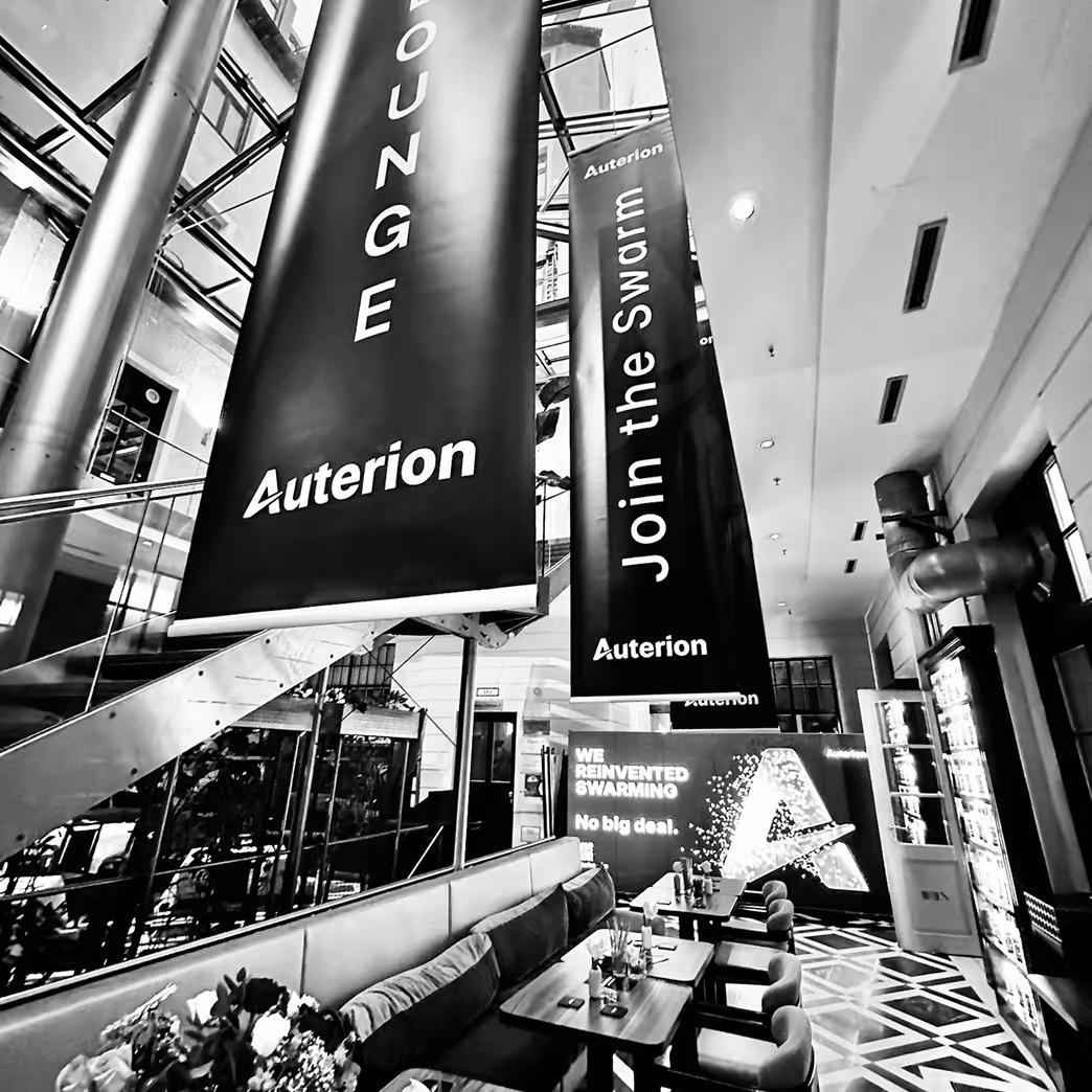 Modern indoor lounge area with hanging banners displaying 'Auterion' and 'Join the Swarm,' featuring tables, chairs, and a lit promotional display.