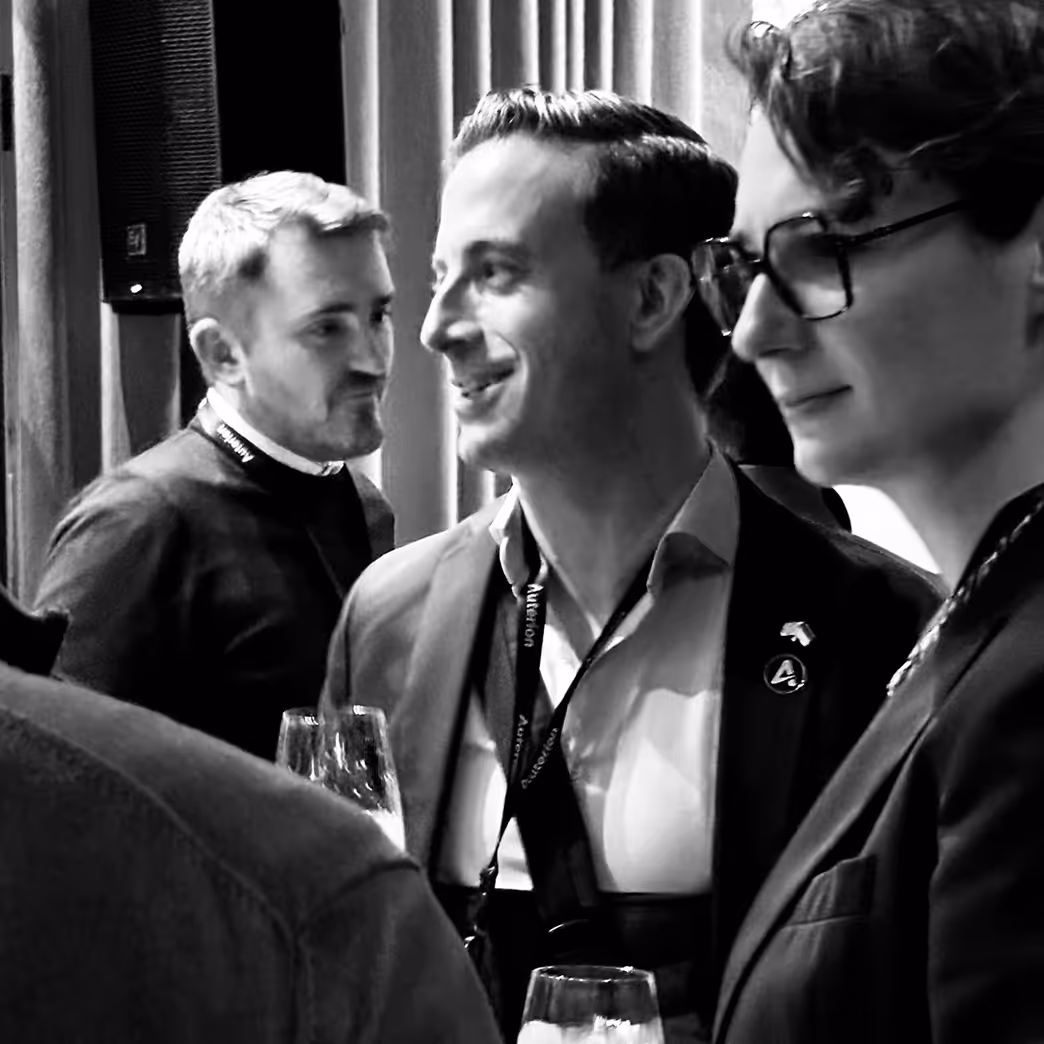 Three men in conversation at a formal event holding wine glasses, one wearing glasses and another with a lanyard and suit.