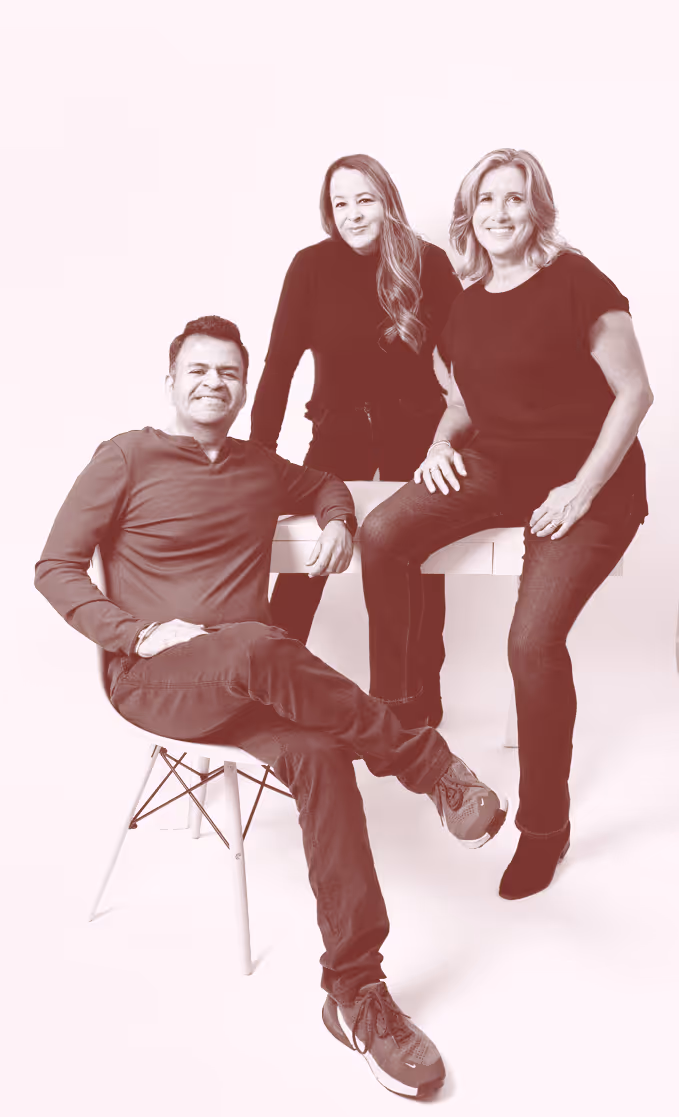 Three people smiling: a man sitting on a chair with legs crossed and two women standing behind a white table.