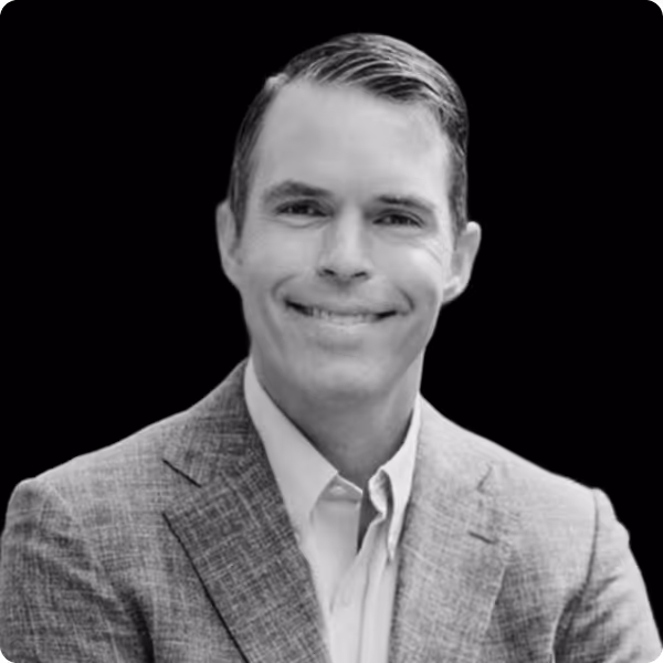 Portrait of a smiling man with neatly combed hair wearing a light-colored shirt and a checked suit jacket against a black background.