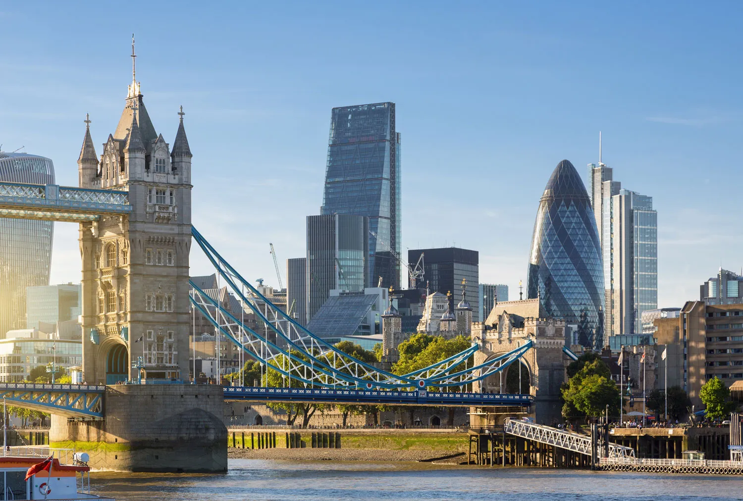 Tower Bridge over the River Thames with London skyscrapers, including the Gherkin and the Leadenhall Building, in the background under a clear sky.