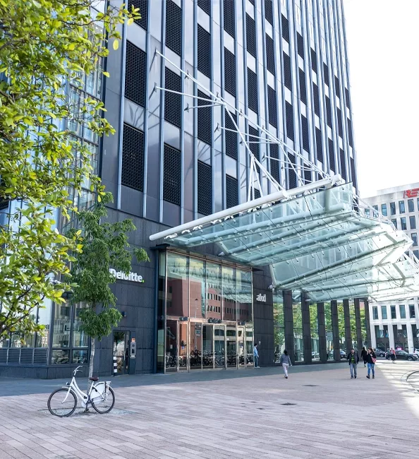 Moderno gebouw met glazen ingang en stalen overkapping in stadscentrum, met fiets en voetgangers op plein.