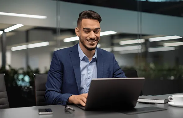 Smiling man in blue suit working on a laptop at a desk in a modern office.