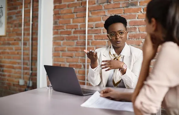 Businesswoman explaining something during a meeting, sitting at a table with a laptop and a document.