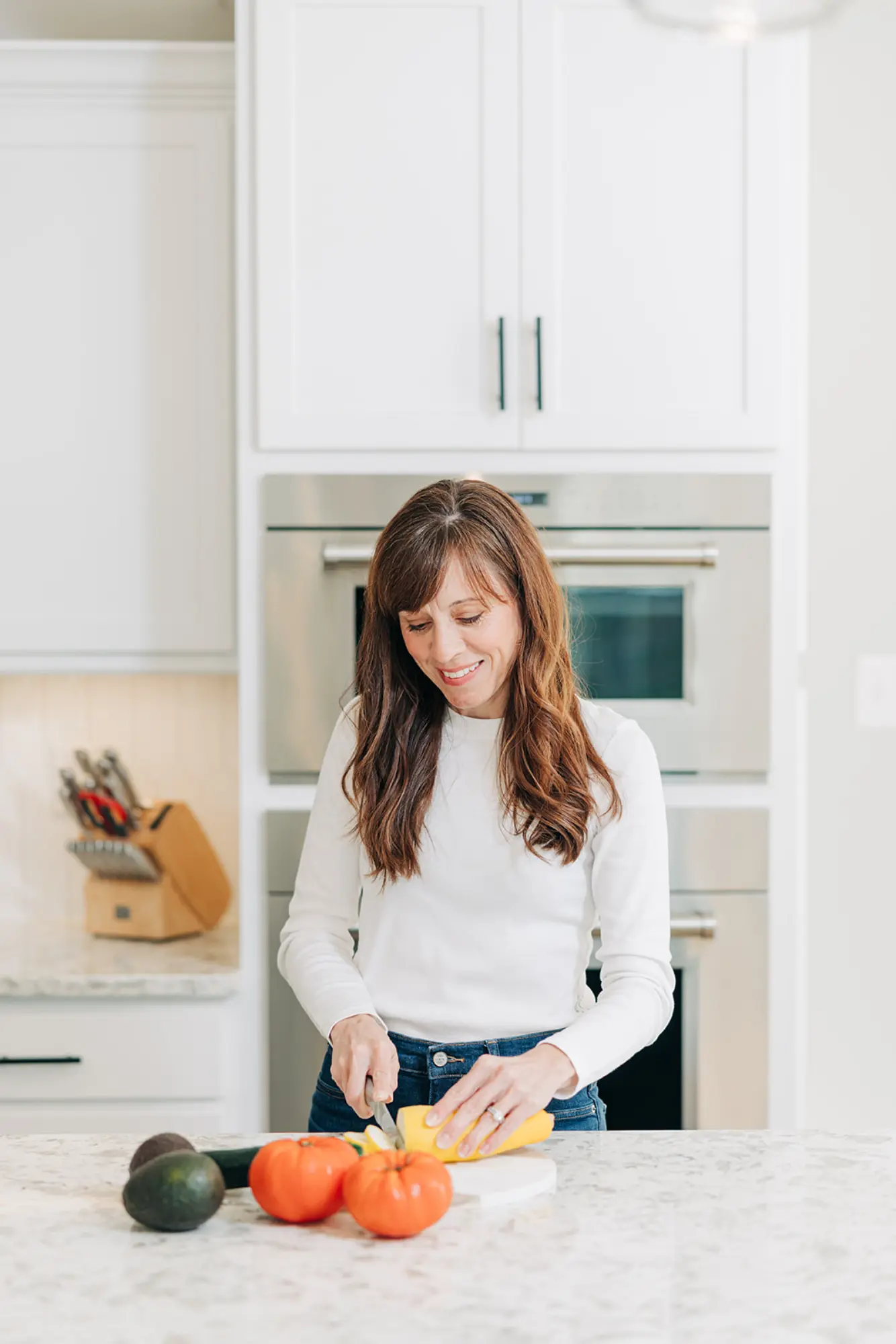 Smiling woman in white shirt slicing yellow squash on a kitchen countertop with tomatoes, avocados, and zucchini nearby.
