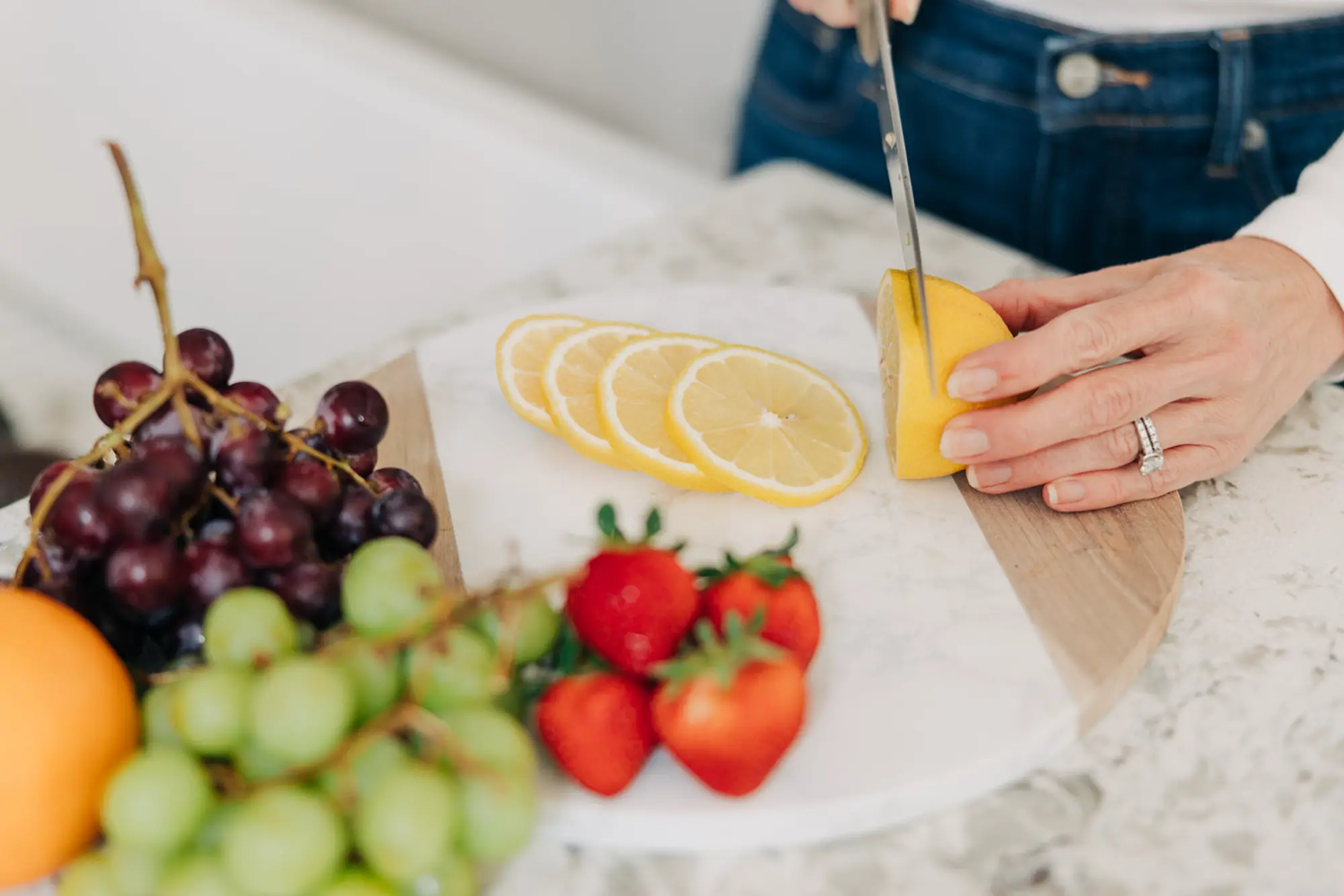 Person slicing a lemon on a round cutting board with strawberries, green and red grapes nearby.