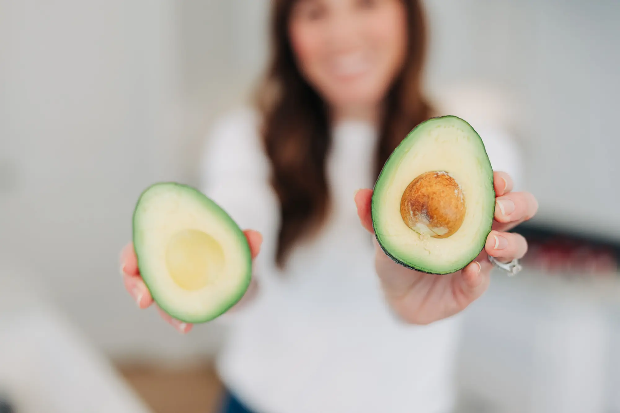A woman holding two halves of a ripe avocado, one with the seed and one without.
