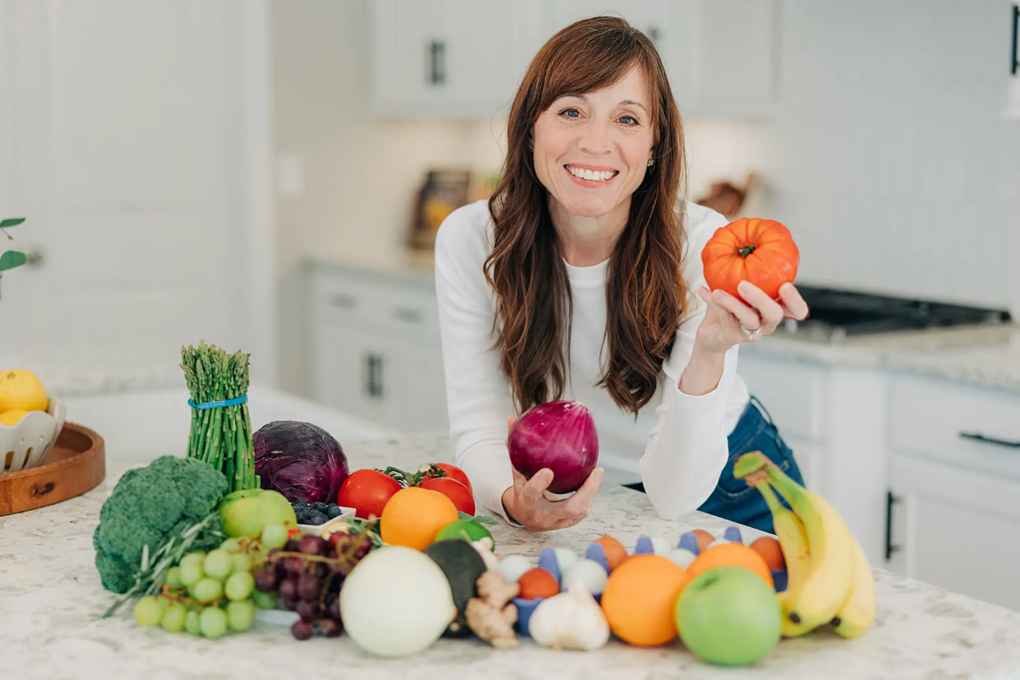 Smiling woman in a white shirt holding a tomato and an onion, standing behind a kitchen counter with assorted fresh fruits and vegetables.