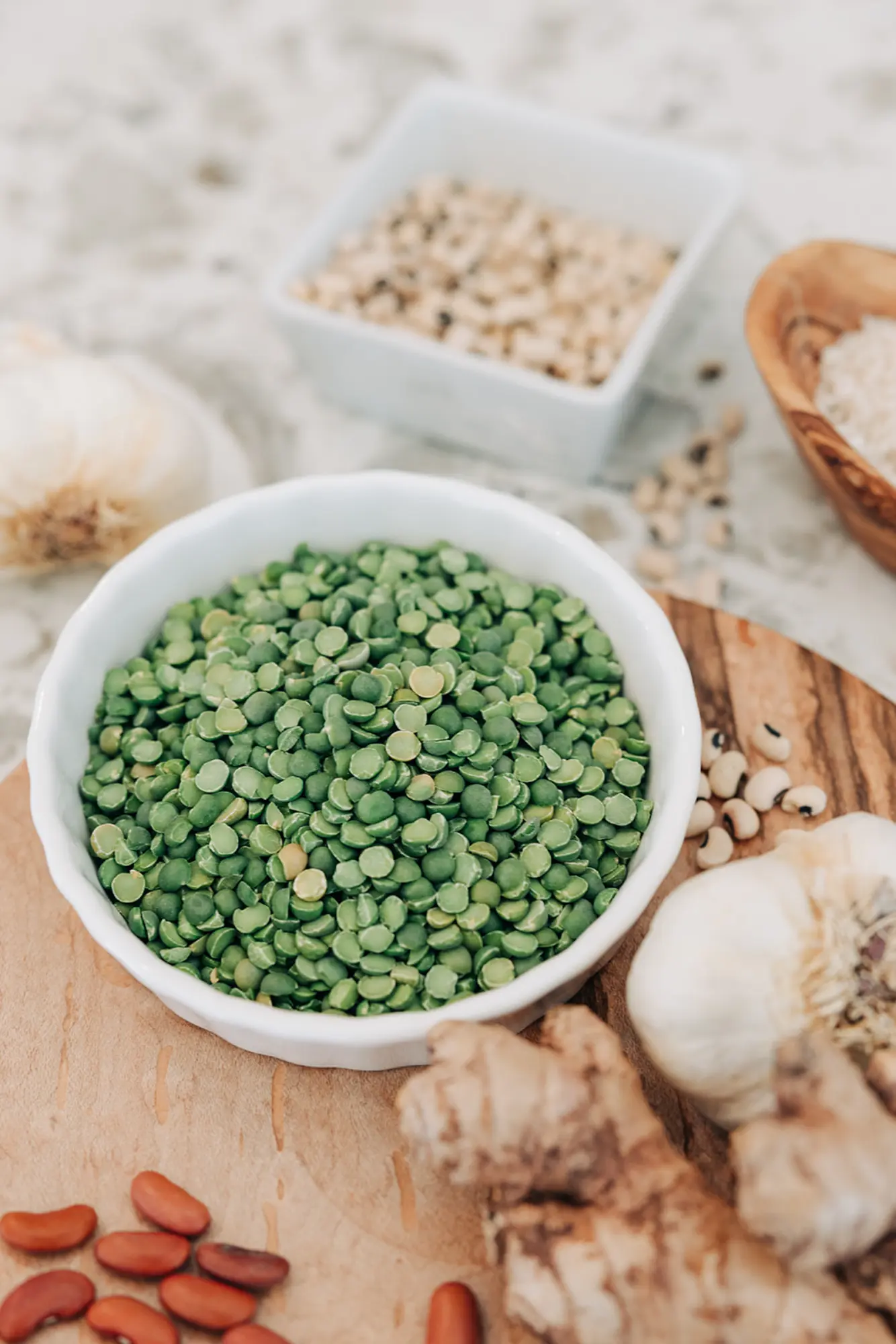 White bowl of green split peas on a wooden board surrounded by garlic bulbs, ginger root, red kidney beans, black-eyed peas, and a bowl of rice.