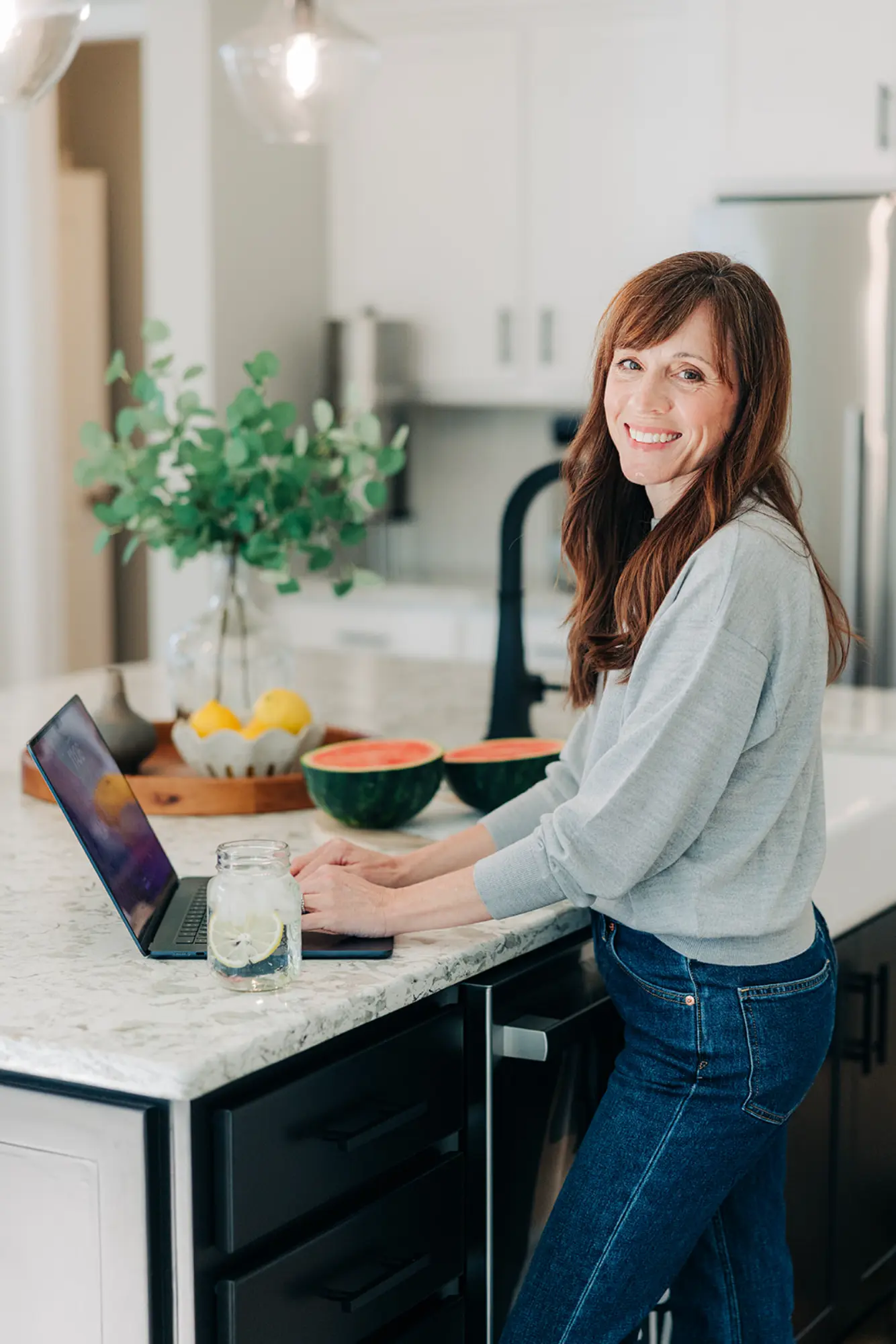 Smiling woman in gray sweater and jeans typing on laptop at kitchen island with watermelon halves and a glass of lemon water.