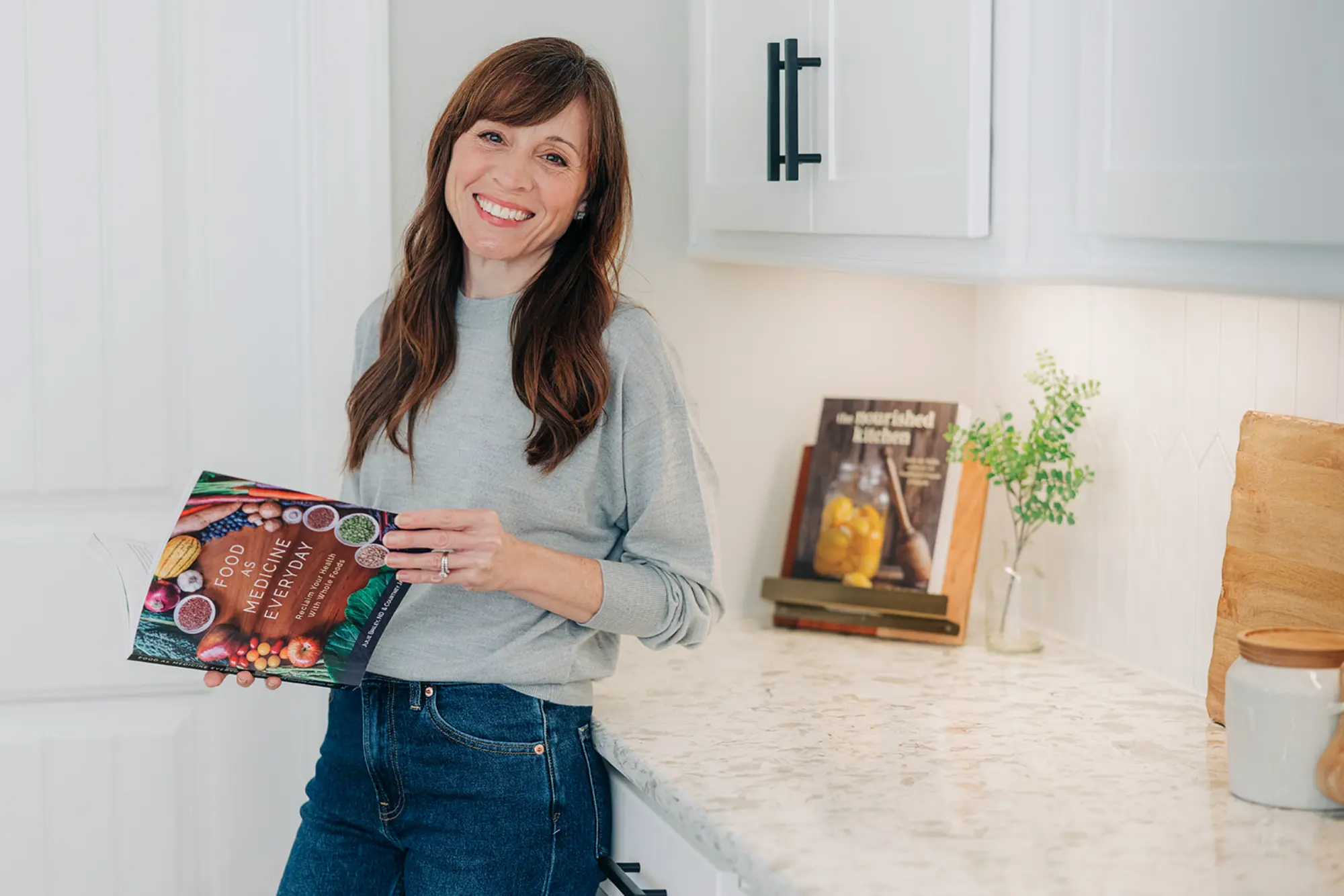 Woman smiling and holding a cookbook titled 'Food as Medicine Everyday' in a bright kitchen.