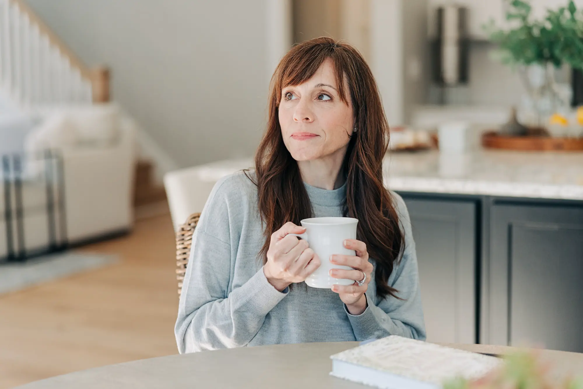 Woman with long brown hair wearing a gray sweater, sitting at a table holding a white mug and looking thoughtfully to the side in a modern kitchen.