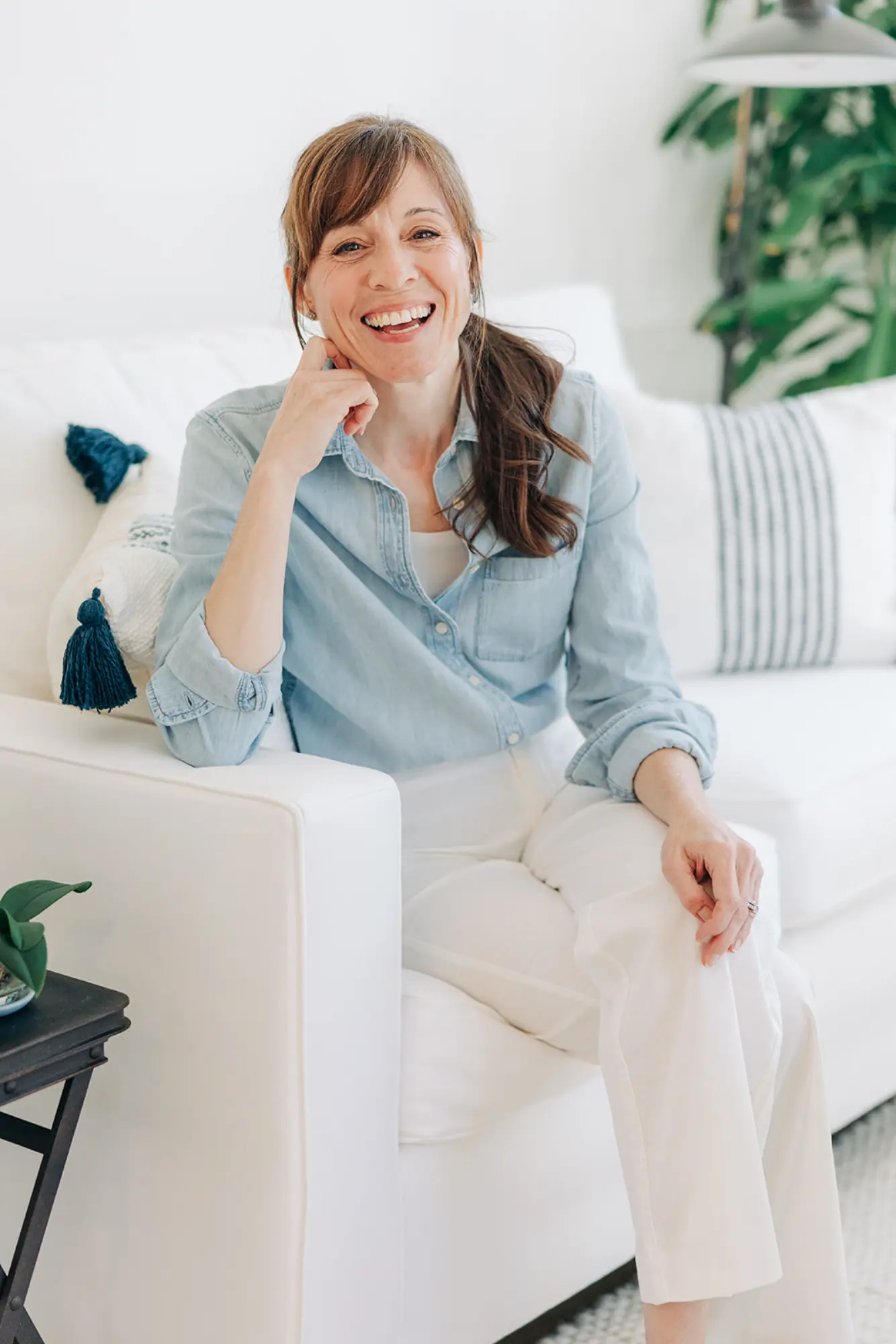 Smiling woman with brown hair in a ponytail wearing a light blue denim shirt and white pants sitting on a white sofa.