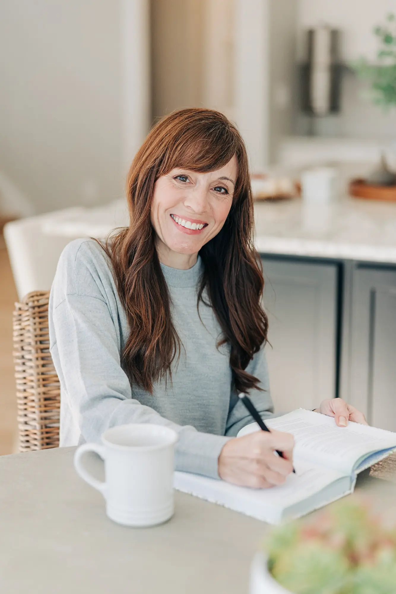 Smiling woman with long brown hair sitting at a table, writing in a book with a white mug in front of her.