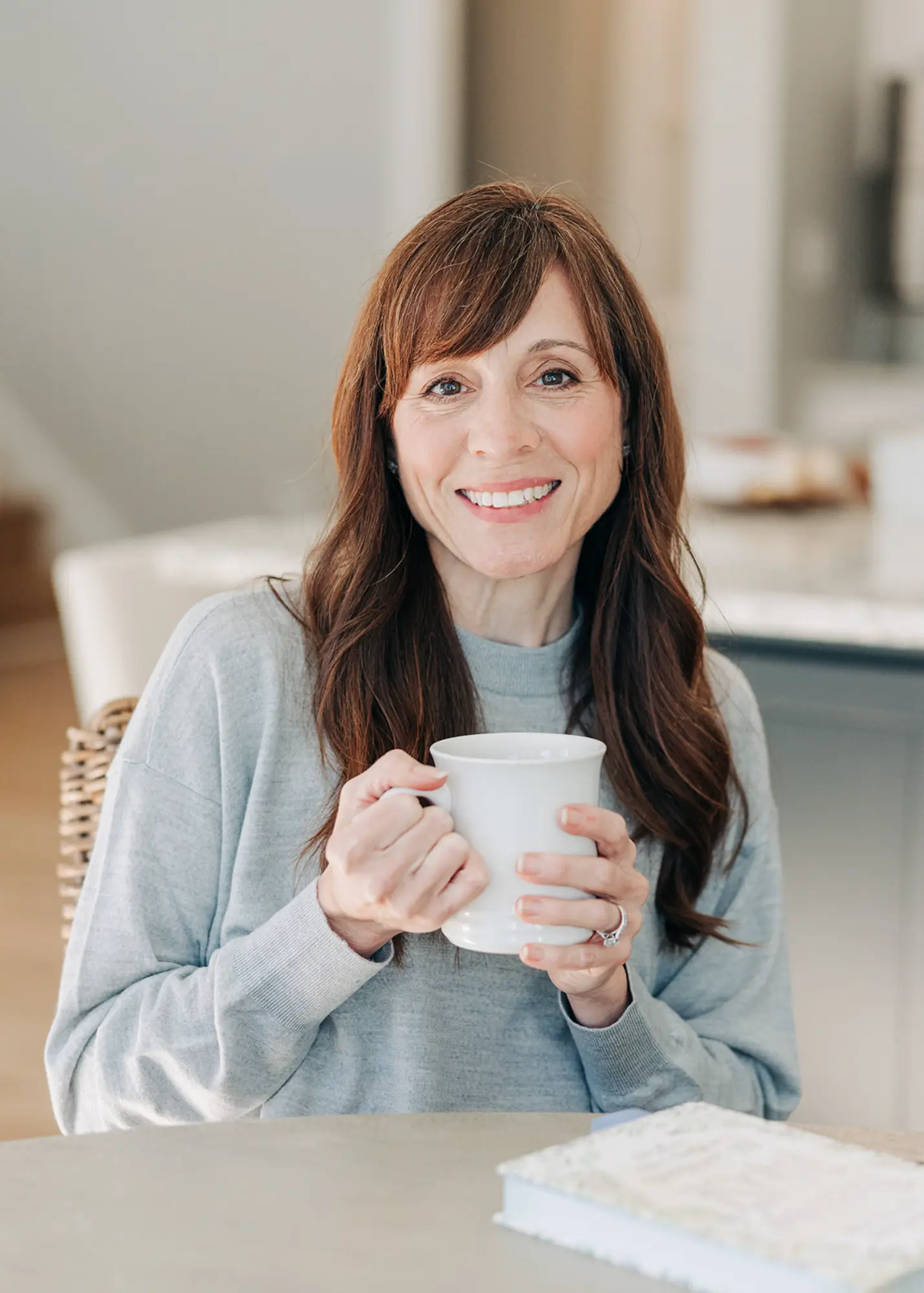 Smiling woman with long brown hair wearing a gray sweater, holding a white mug indoors.