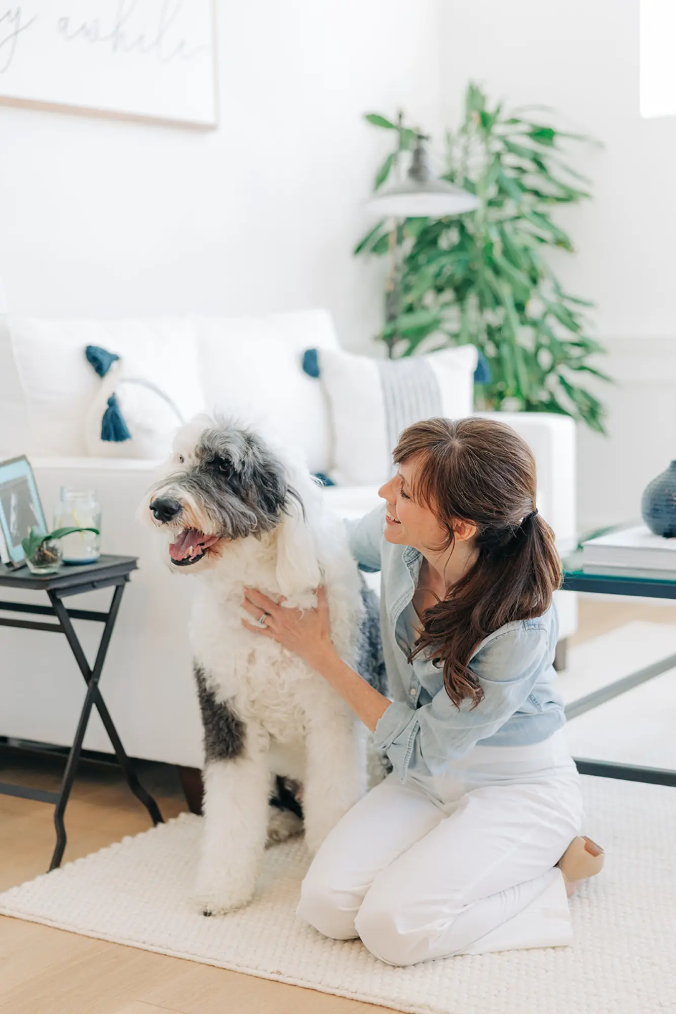 Woman kneeling on a carpet smiling and petting a large fluffy black and white dog in a bright living room.