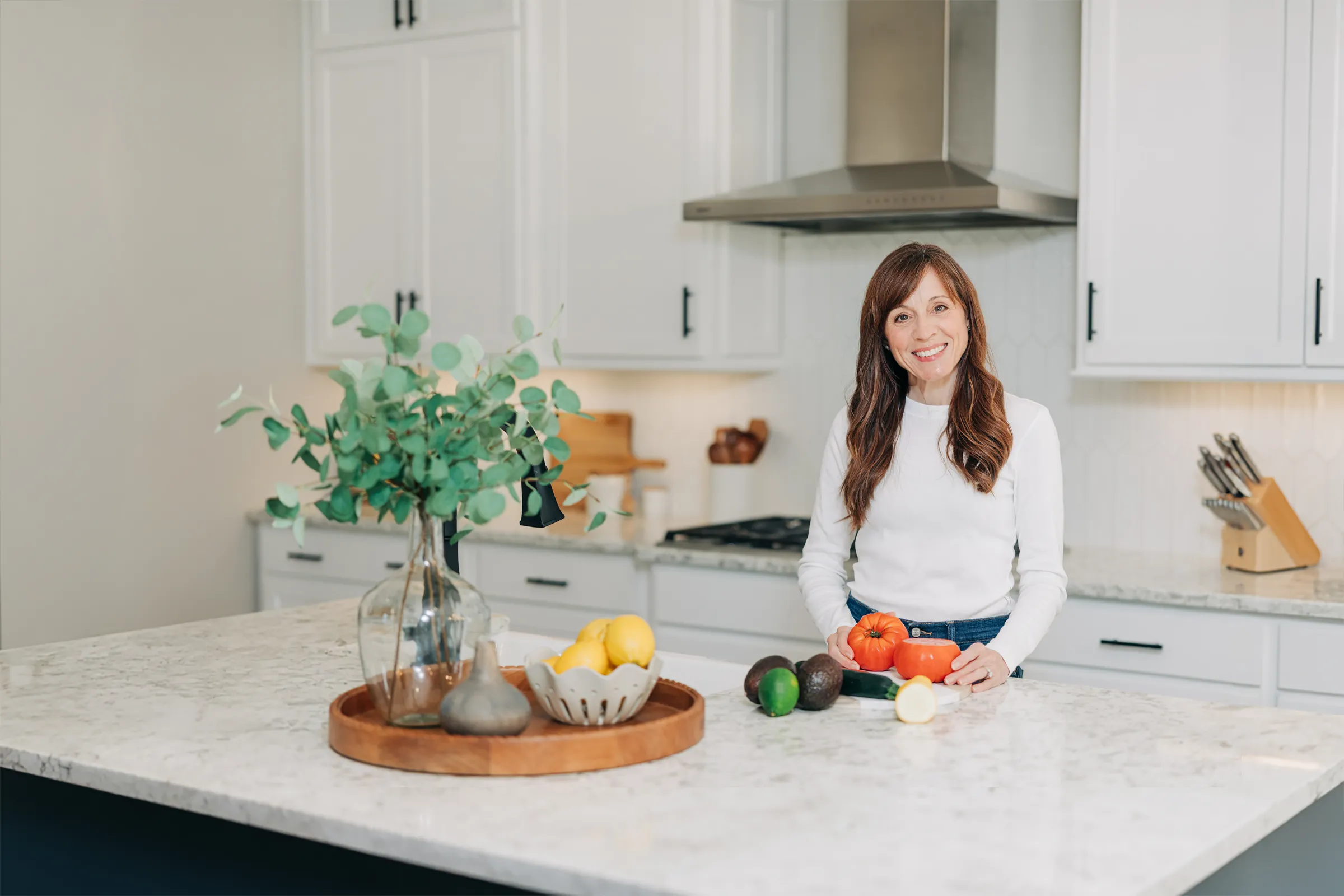Woman smiling in a modern kitchen with fresh vegetables and lemons on a marble countertop.