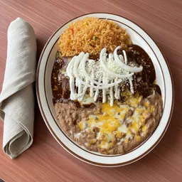 Plate of Mole Poblano with Mexican rice, refried beans with melted cheese, and mole sauce topped with sour cream and sliced onions, next to a rolled white napkin.