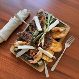 Grilled meat platter with fried plantains, shrimp, zucchini, green chili, and sliced white cheese on a square plate with fork and knife on wooden table.