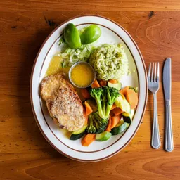 Plate with grilled salmon, green rice, steamed broccoli, carrots, lettuce, two lime wedges, and a small bowl of sauce on a wooden table with fork and knife.