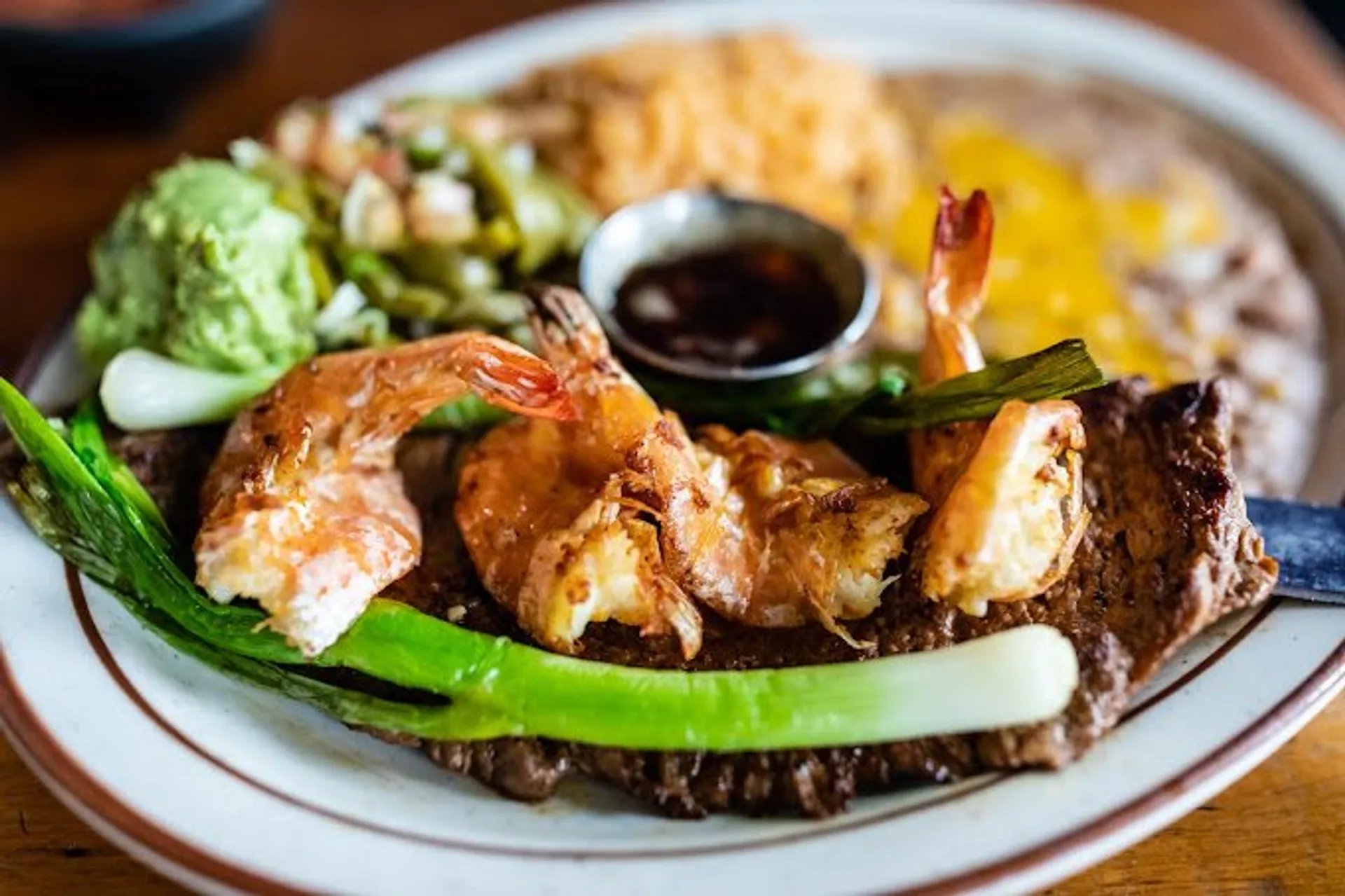 Plate with grilled shrimp on steak, green onions, guacamole, rice, and refried beans with melted cheese.