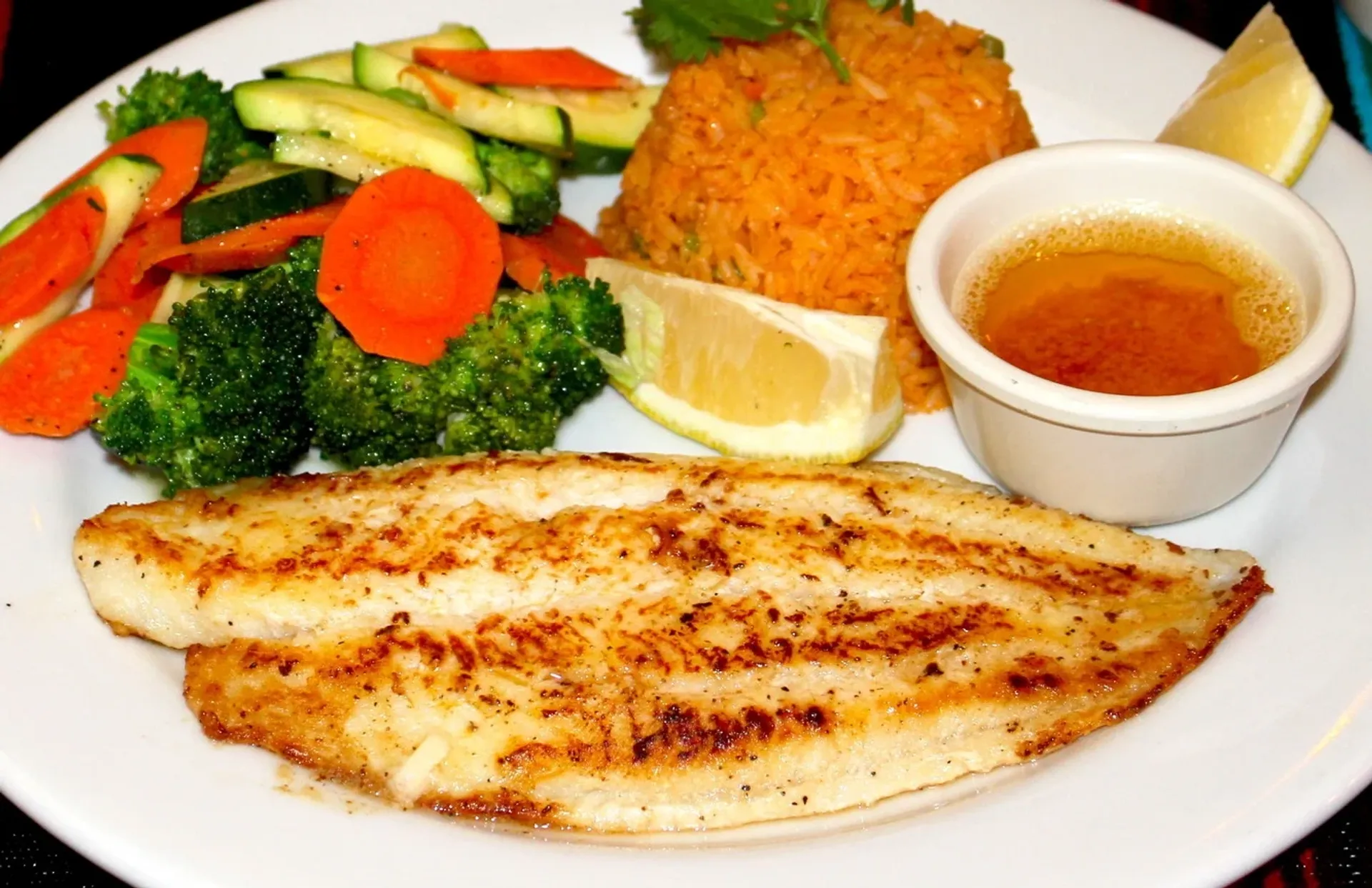 Grilled fish fillet served with mixed steamed vegetables, seasoned rice, lemon wedges, and a small cup of dipping sauce on a white plate.