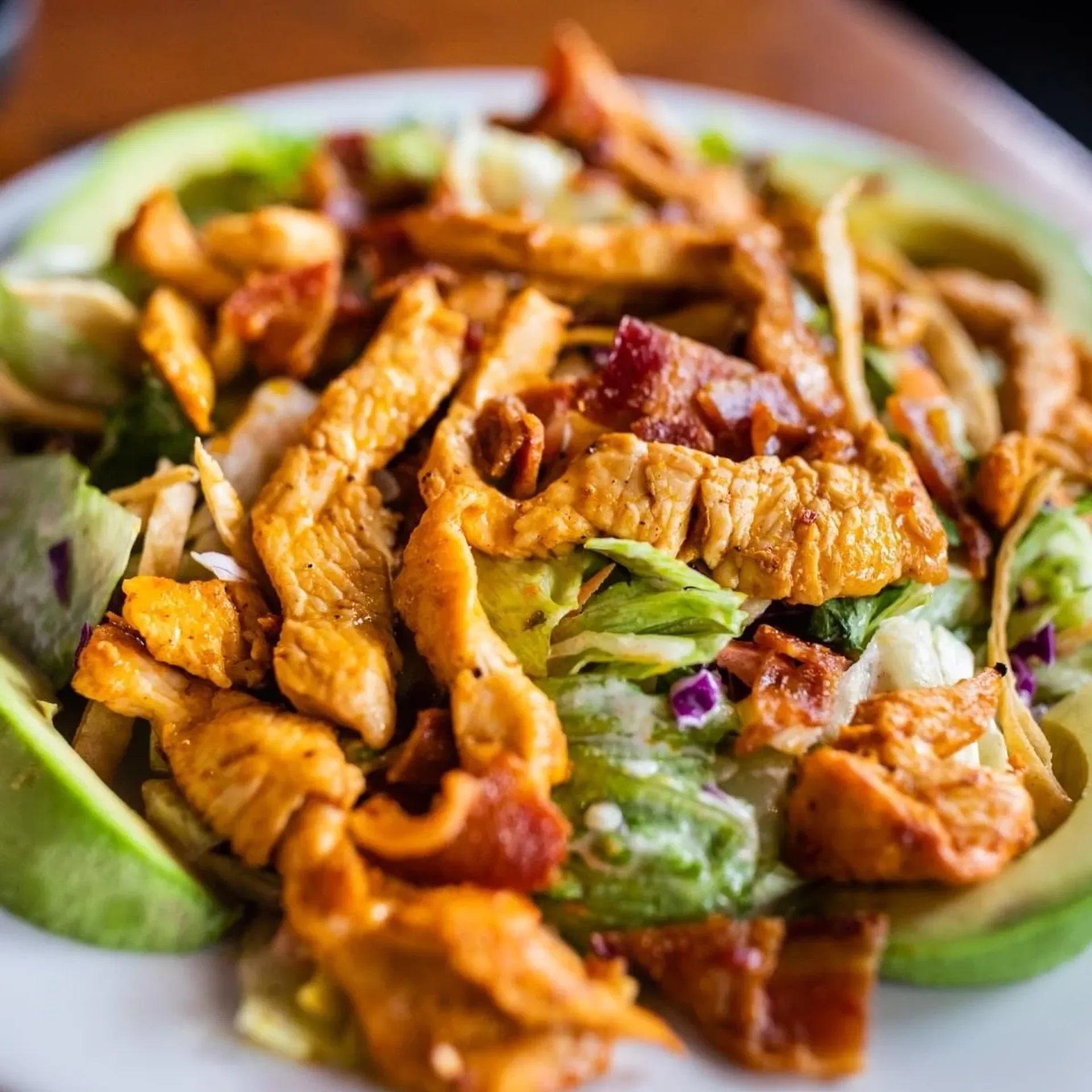 Close-up of a salad topped with grilled chicken strips, bacon pieces, lettuce, and avocado slices.