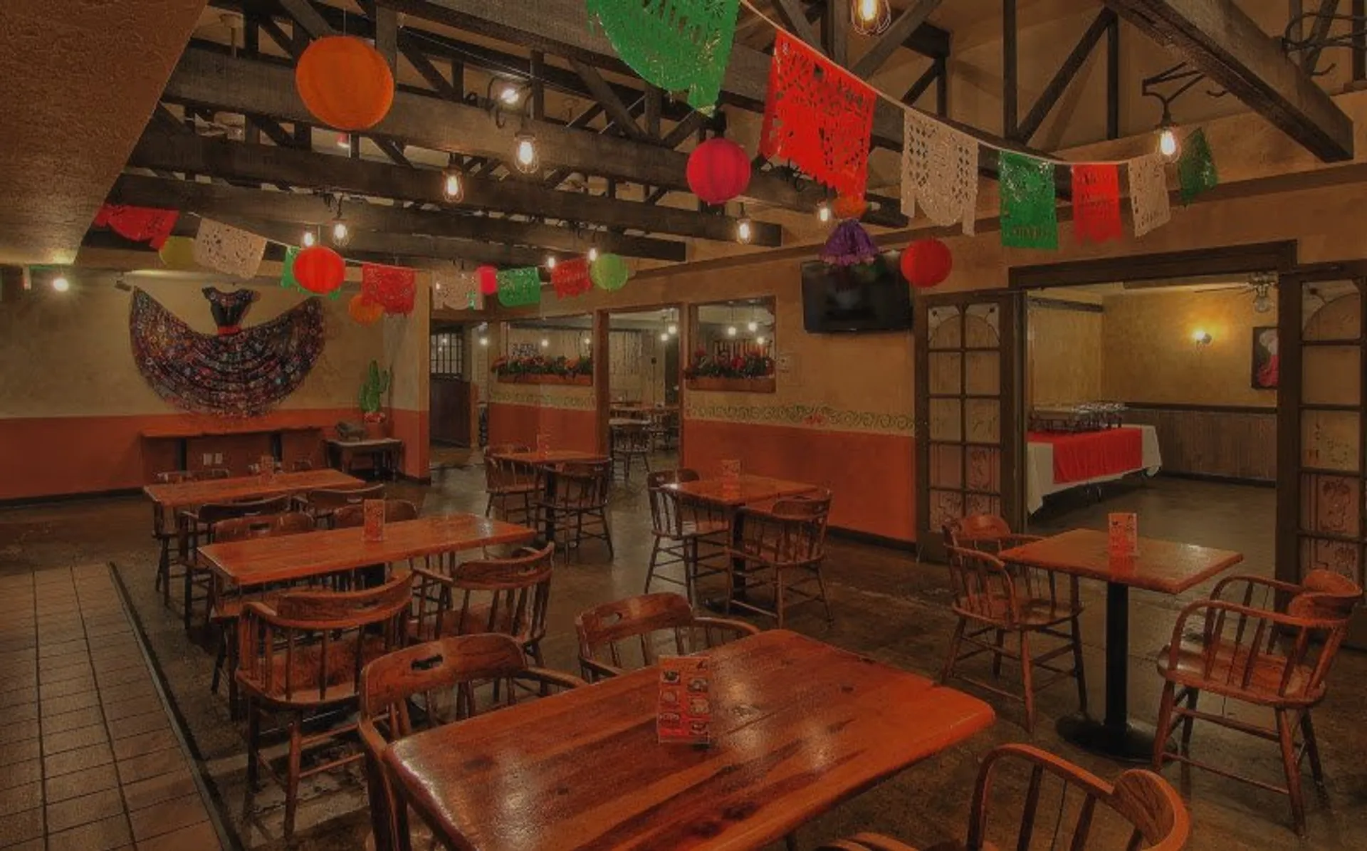 Interior of a Mexican restaurant with wooden tables and chairs, colorful papel picado decorations hanging from the ceiling, and a decorative wall hanging resembling a traditional Mexican skirt.