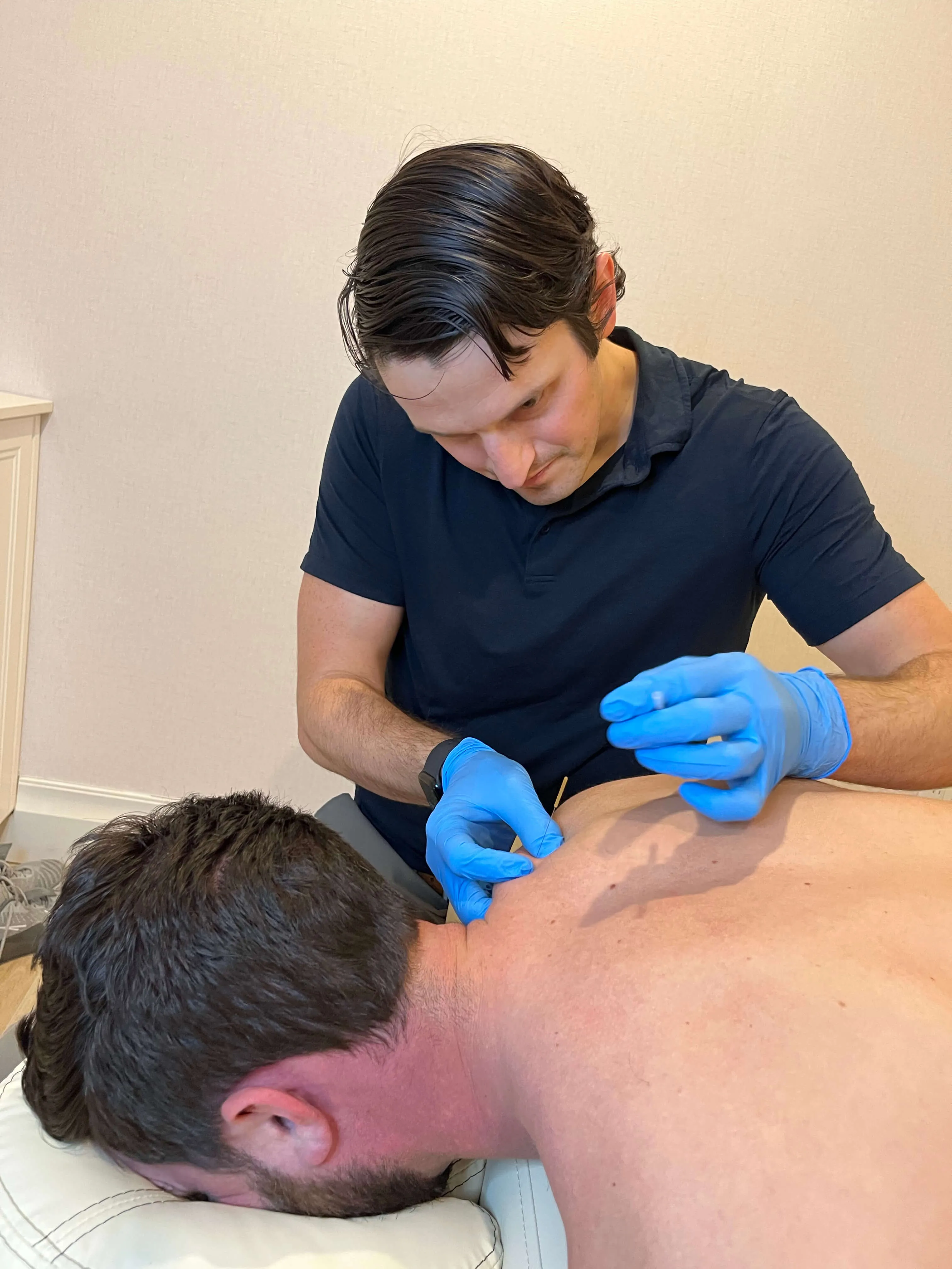 A clinician wearing blue gloves performing dry needling on a man's upper back and neck area while he lies face down on a treatment table.