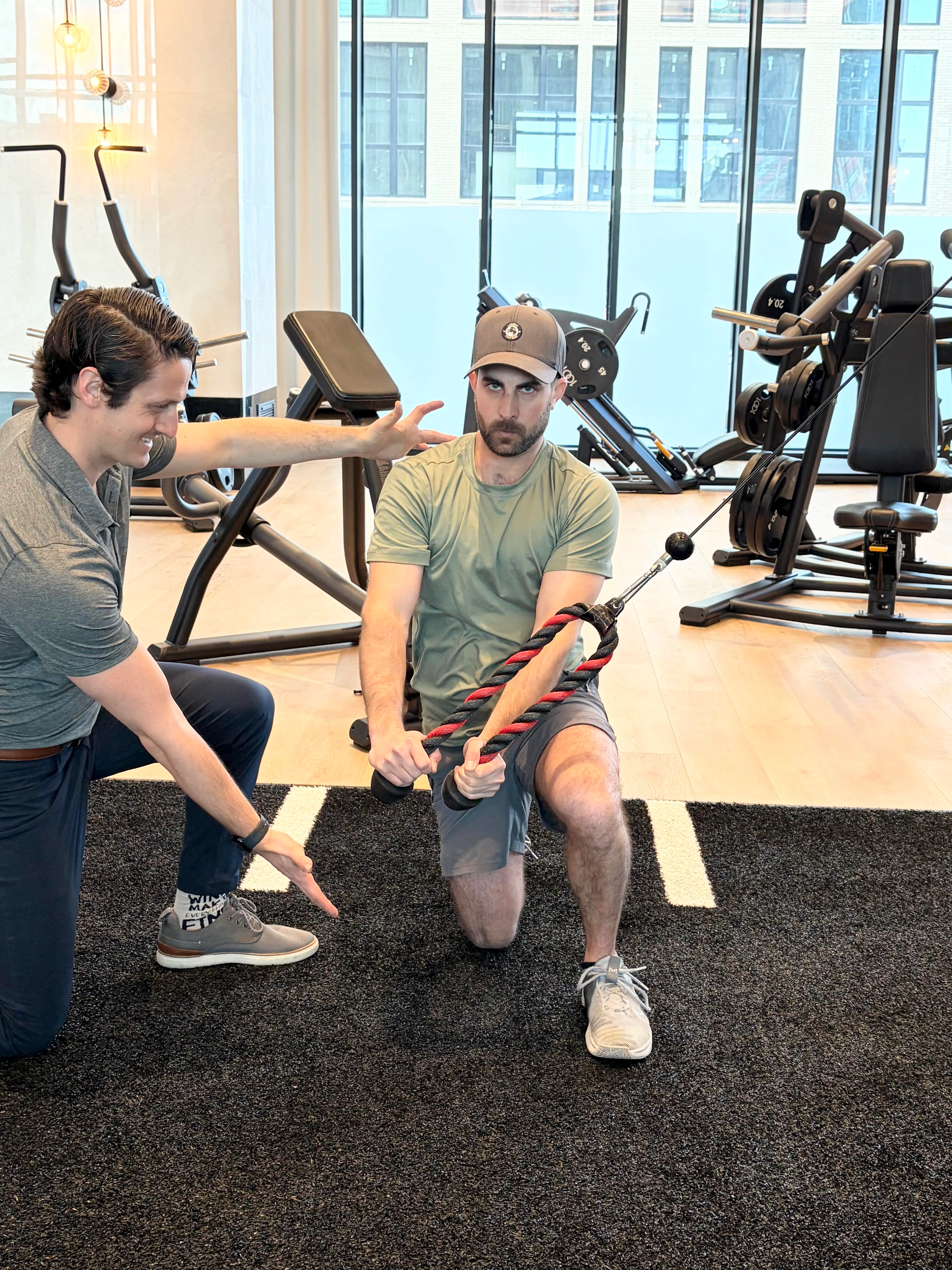 Man in a green shirt and cap performing a half-kneeling cable chop exercise with a trainer guiding him in a gym.