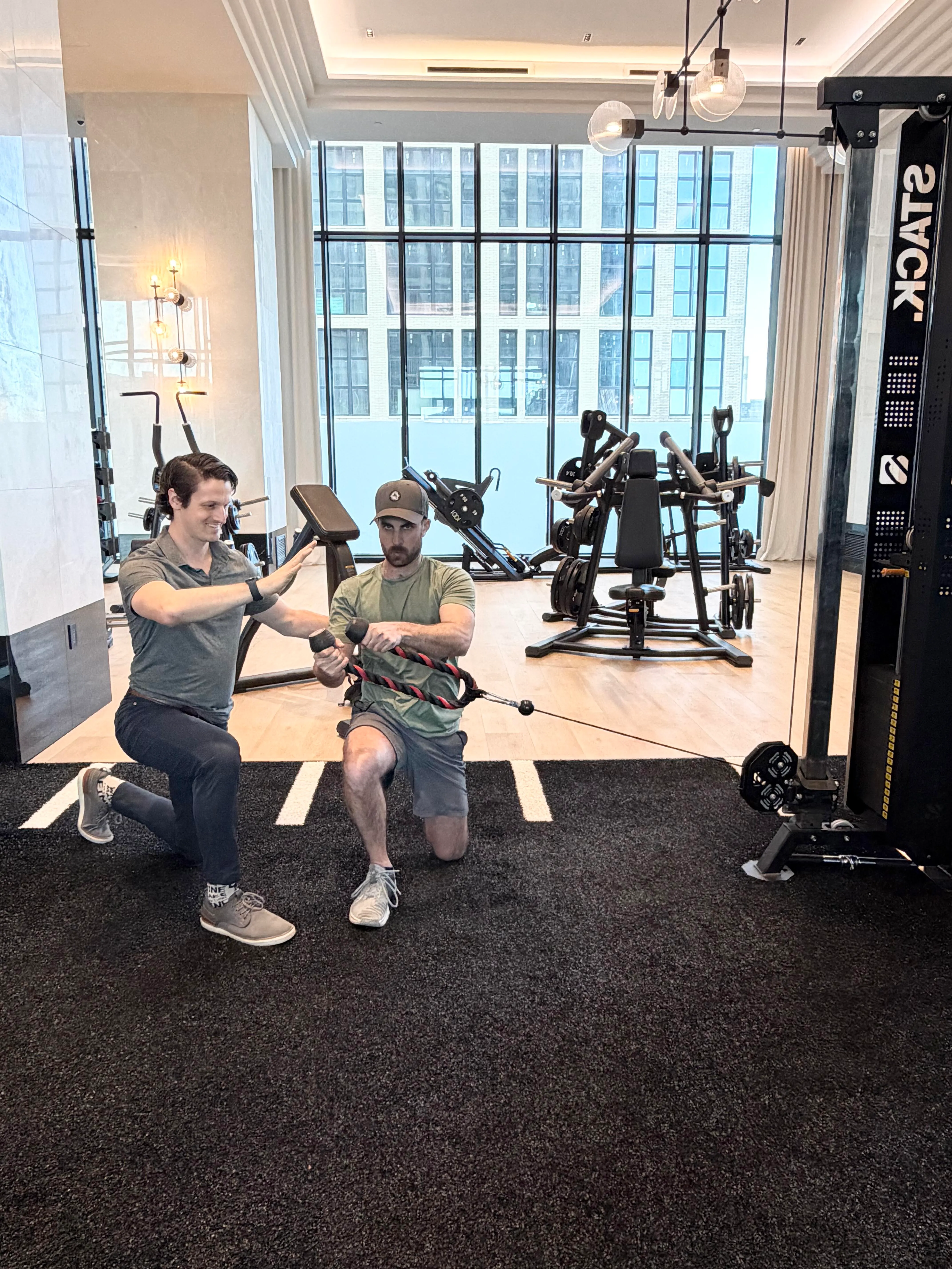 Man in green shirt performing half-kneeling cable row exercise with resistance rope, guided by trainer in gym.