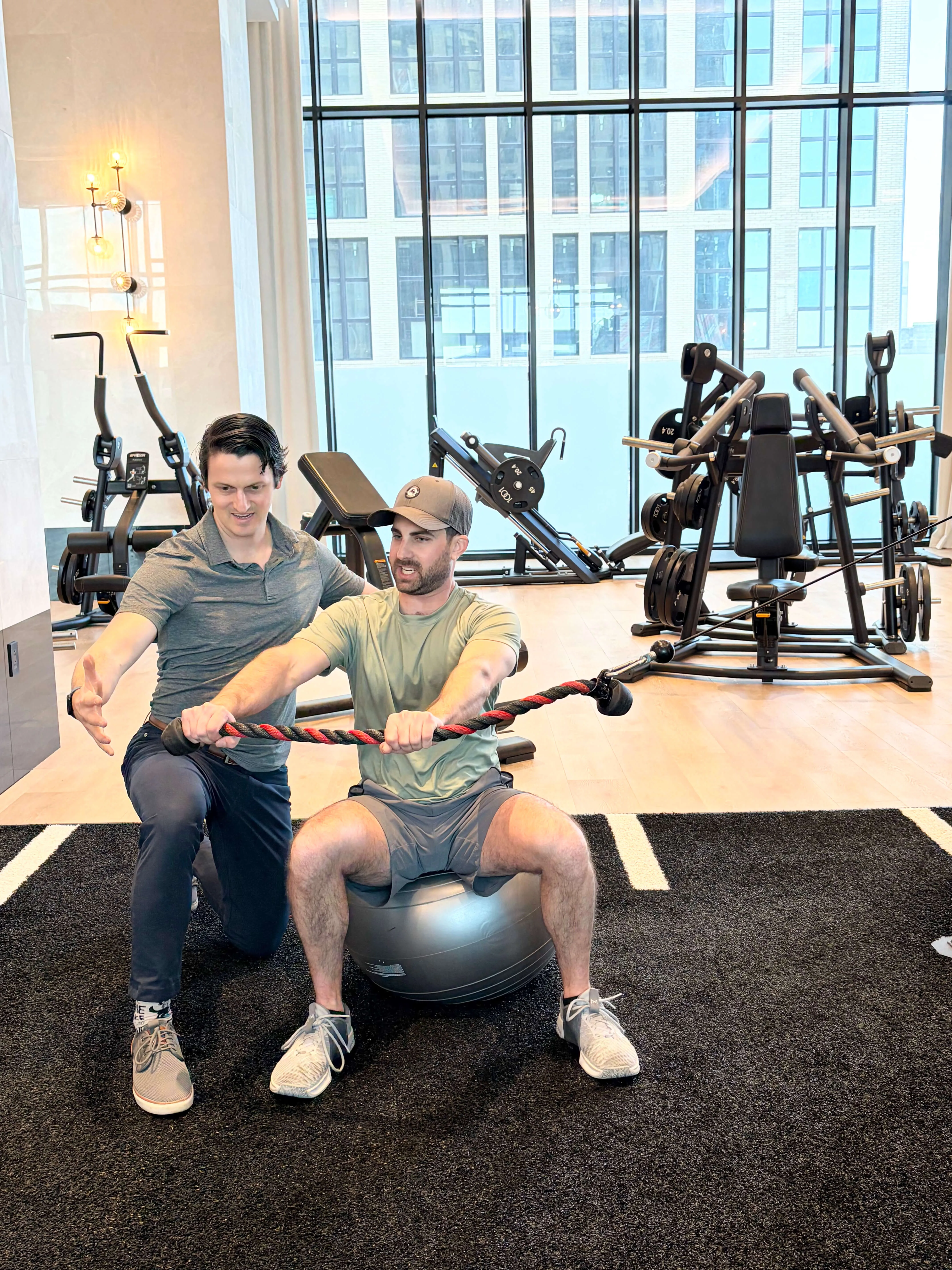 Man sitting on exercise ball holding a rope performing seated cable rotation exercise while trainer kneels beside him giving guidance in a gym.