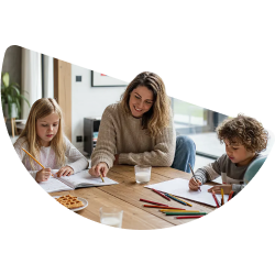 Een glimlachende vrouw helpt twee kinderen met tekenen met kleurpotloden aan een houten tafel.