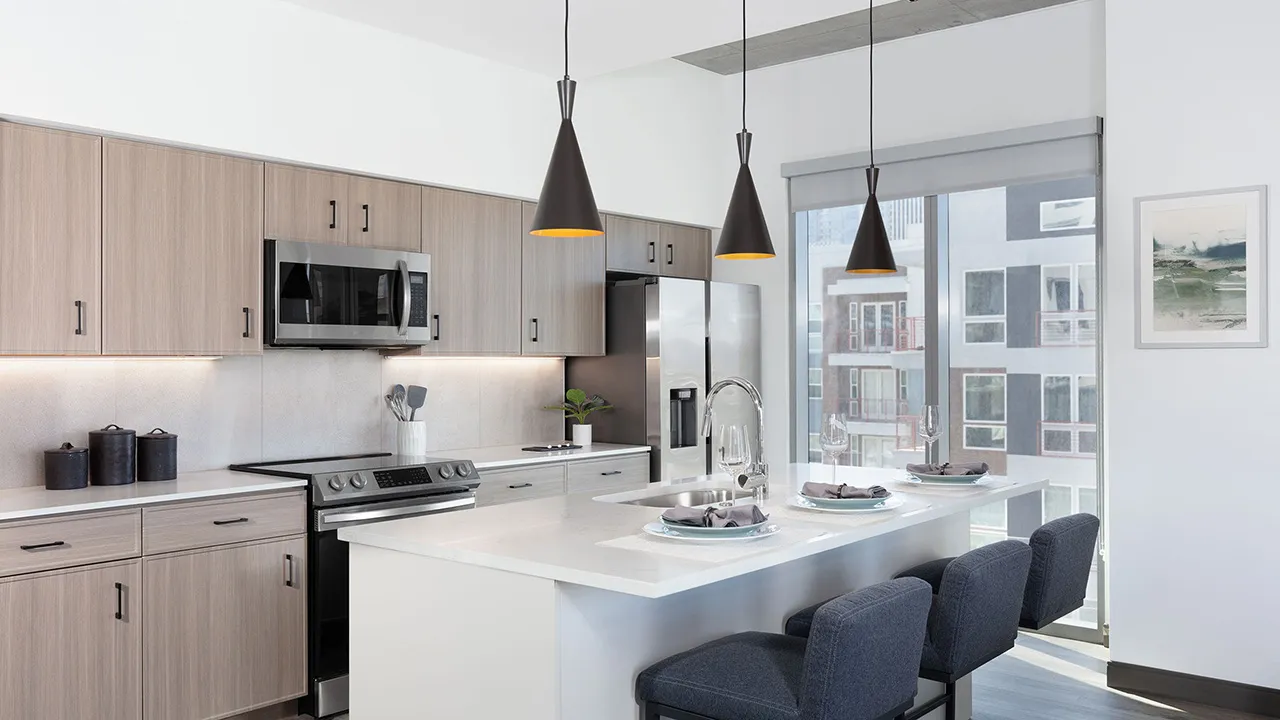 Modern kitchen with light wood cabinets, stainless appliances, island with sink and three dark chairs.