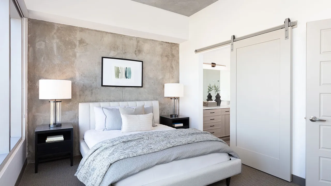 Modern bedroom with white bed, blue-gray bedding, nightstands with lamps, and sliding bathroom door.