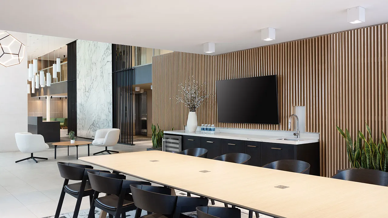 Modern meeting room with long table, black chairs, wall-mounted TV, and wooden slat wall with plants.