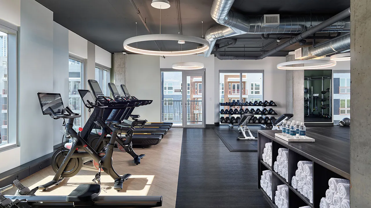 Modern gym with exercise bikes, treadmills, dumbbells rack, benches, rolled towels, and water bottles.